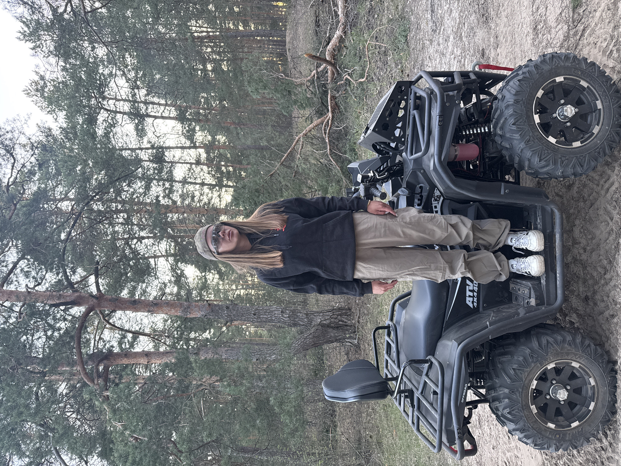A person is standing inside a black ATV in a wooded area. The ATV has large, knobby tires and a rear cargo rack. The environment appears to be a forest near sandy ground. It could be near Bol'shoye Kozino, Russia, as the location was included in the prompt, so it may be a forest or park area in that vicinity. The person is wearing a dark hoodie, light-colored pants, and sneakers. They also have a beanie and sunglasses.