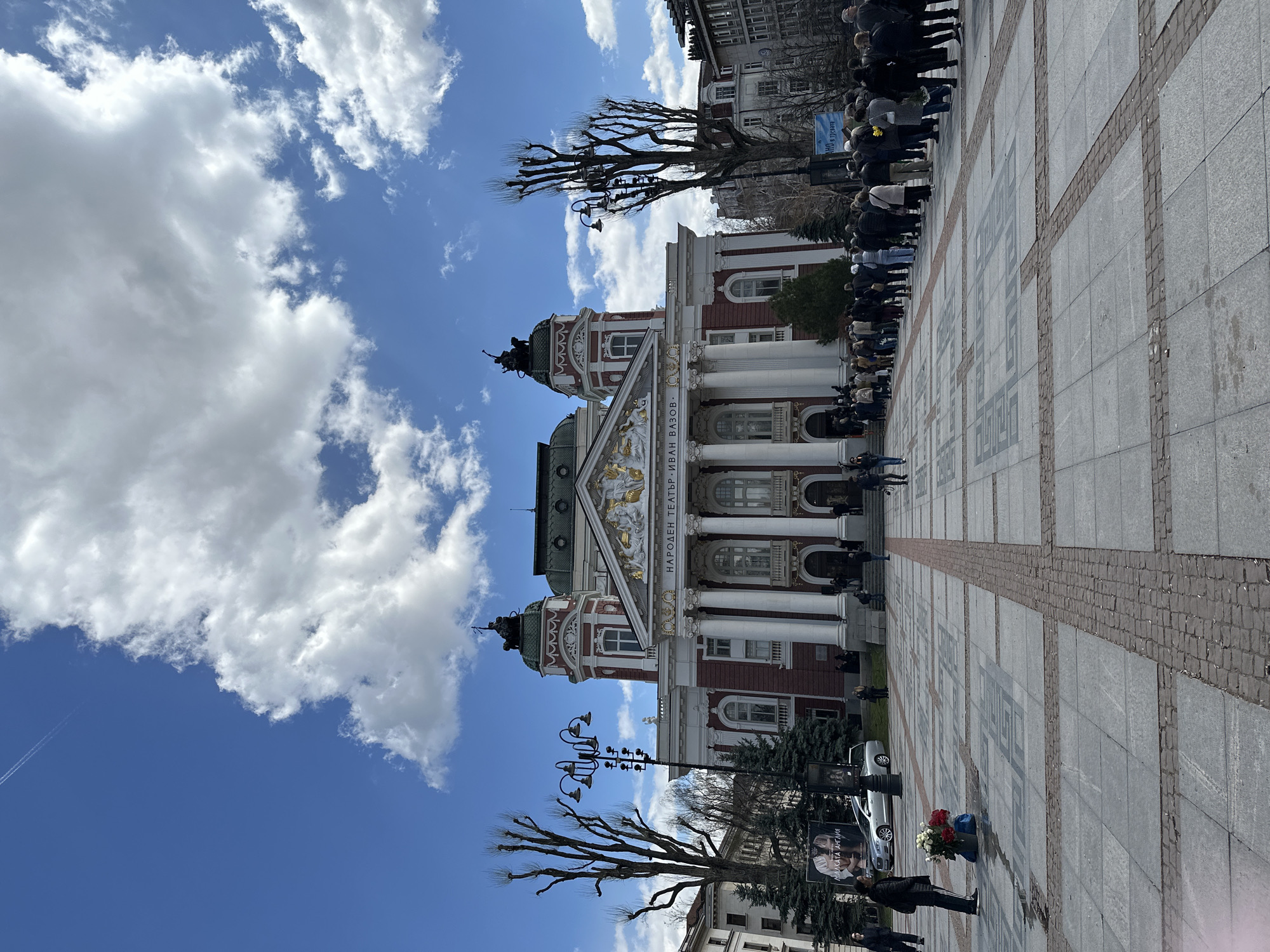The image shows the Ivan Vazov National Theatre in Sofia, Bulgaria, on a sunny day with a partly cloudy blue sky. The theatre's facade is visible, with its white columns and reddish-brown brickwork, along with the gold-colored sculptures on the pediment. There is a large crowd of people gathered near the building. Some are standing in line, and others are in groups. A few trees with bare branches are in the foreground. On the right side of the image, the ground is paved with gray and brown stones. A bouquet of flowers in a bucket stands on the paved area.