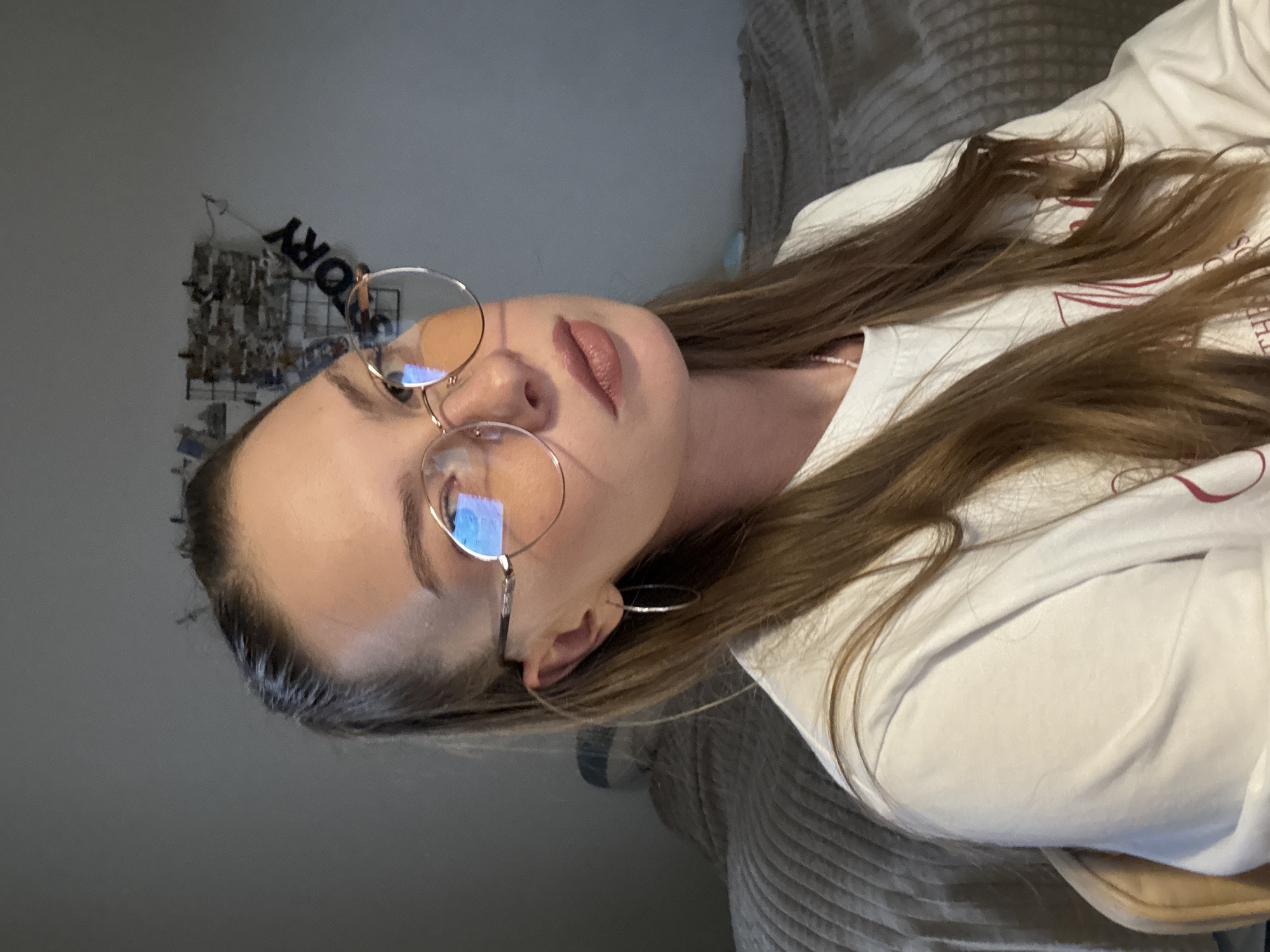 This is a media file showing a close-up, vertically oriented shot of a young woman with long, brown hair and light-colored eyes wearing large, round glasses. She has on a white t-shirt. The background appears to be an indoor setting with a wall and a bed. The lighting suggests it is evening or night, with a single light source casting shadows and creating reflections in her glasses. There is a wall-mounted decorative item behind her with the letters "ORY" visible, possibly part of a larger word or phrase. The woman's facial expression is neutral. The scene has a relaxed and casual feel.