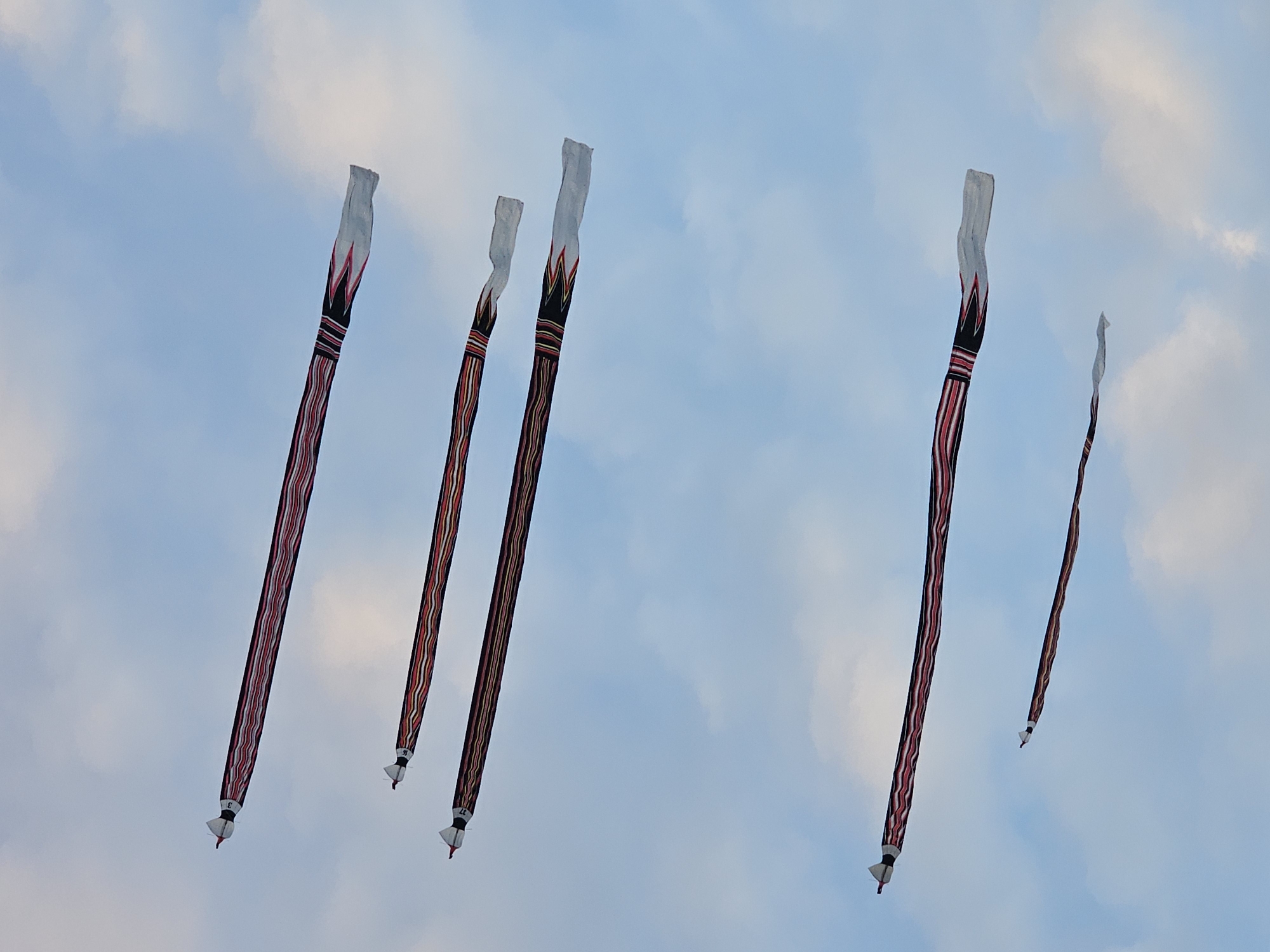 Five traditional Balinese kites soar in the sky over Denpasar, Indonesia. The kites are long and narrow, with white heads decorated with red and black patterns. Their bodies are striped with red, black, and brown. Small white tassels hang from their tails. The sky is light blue with wispy white clouds.