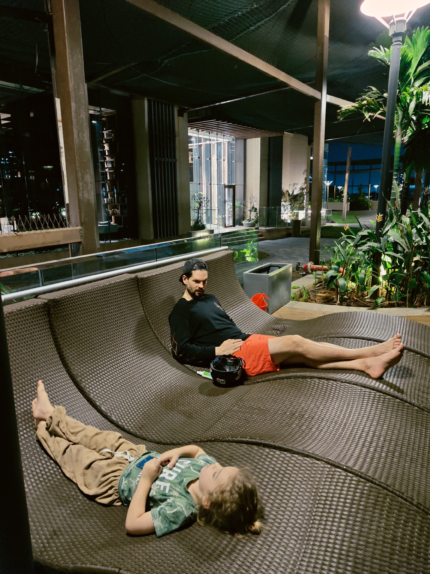 The image depicts a tranquil nighttime scene in an outdoor lounge area, likely part of a modern building complex in Mumbai, India.
**Subjects:**
Two individuals are present: an adult male and a child. The adult male, with dark hair and a beard, is lying on a large, dark brown, woven outdoor lounger. He wears a black long-sleeved shirt and orange shorts, with his bare feet extended. A black helmet and a small white bottle are placed next to him on the lounger. Below him, a child with light-colored hair is also reclining on the lounger, wearing a green patterned t-shirt that partially shows the word "LIVE" and light brown pants. The child's hands rest on their stomach, and their bare feet are visible.
**Scene and Setting:**
The setting is an elegantly designed outdoor leisure space, characterized by modern architecture. The background features tall, contemporary buildings with glass facades and illuminated interiors, set against the dark night sky. Lush tropical-like foliage, including palm trees and various green plants, adds to the serene ambiance. The area is lit by artificial light sources, including a prominent outdoor lamp with a white diffuser in the upper right, and subtle lighting illuminating the plants and pathways. A metal railing with glass panels runs along the edge of the lounging area, separating it from other sections of the complex. Overhead, a dark safety net (or mesh structure) spans the space. Other objects in the background include concrete pillars, a square grey planter, and a red bucket.
**Activity and Emotions:**
Both the adult and the child are engaged in a relaxed activity, resting comfortably on the contoured lounger. The adult appears to be observing his surroundings, while the child seems to be resting or sleeping peacefully. The overall mood conveyed is one of relaxation, calm, and tranquility.
**Time of Day:**
The image is clearly taken at night, indicated by the darkness, the reliance on artificial lighting, and the illuminated cityscape in the distance.