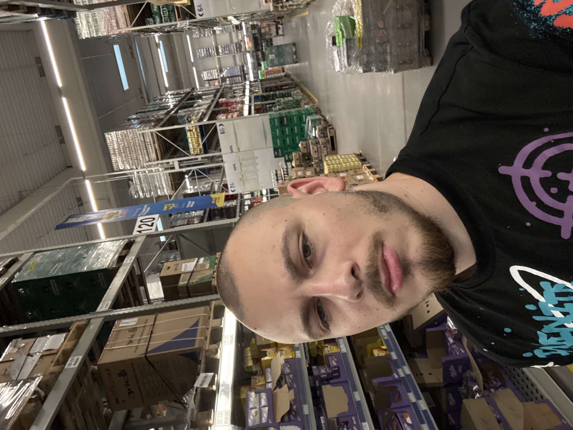 A man with short hair and a beard is taking a selfie in a warehouse-style store with high shelves stocked with goods. The shelves are labeled with numbers, including "120," "320," and "321." Some labels indicate "БИРА" (beer in Bulgarian). Based on these clues, the location is likely a wholesale store, possibly in Stara Zagora, Bulgaria.