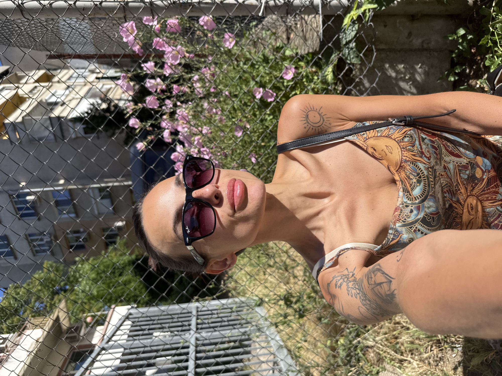In Pleven, Bulgaria, a woman with her hair pulled back, wearing sunglasses and a patterned tank top, takes a selfie outdoors. She has tattoos on her left arm and shoulder. Behind her, a chain-link fence separates her from a backdrop of buildings and lush green trees, with a cluster of pink flowers in the mid-ground. The sun casts strong shadows, indicating a bright day.