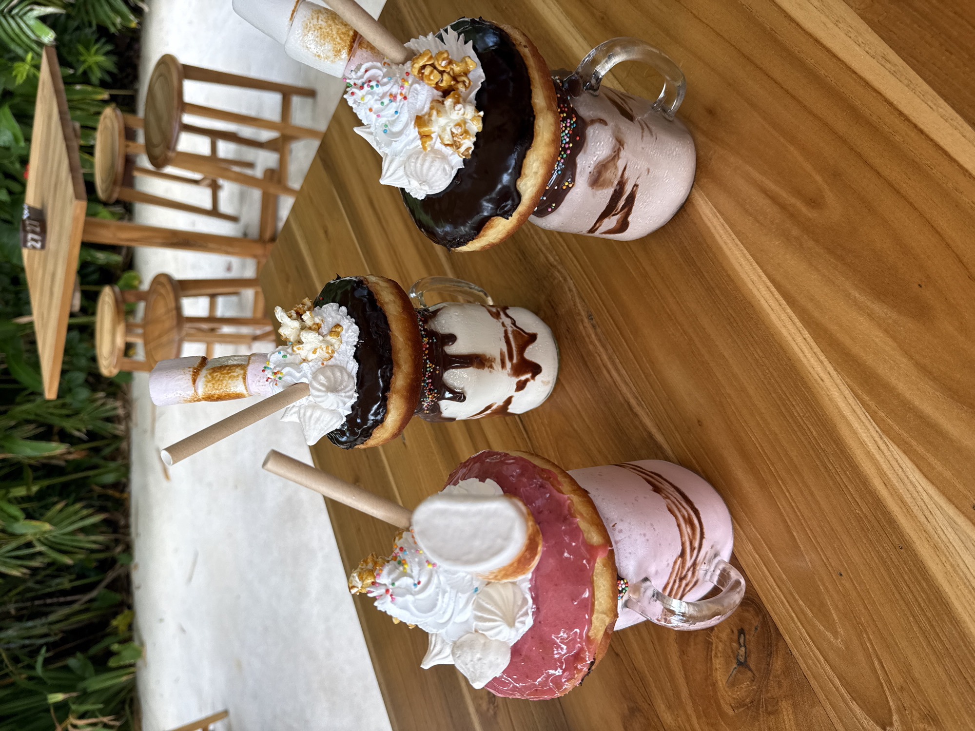 The image shows three elaborately decorated milkshakes on a wooden table. Each milkshake is served in a glass mug with a handle and is topped with a doughnut, whipped cream, popcorn, sprinkles, and a toasted marshmallow on a stick. The doughnuts are glazed in different colors: chocolate, vanilla, and strawberry. The milkshakes themselves are also different colors, corresponding to the doughnut glaze colors.
In the background, there are several wooden stools, and some green foliage is visible. The image appears to have been taken at a cafe or restaurant, likely in Kuta, Indonesia, given the tropical vegetation and the style of the food presentation.