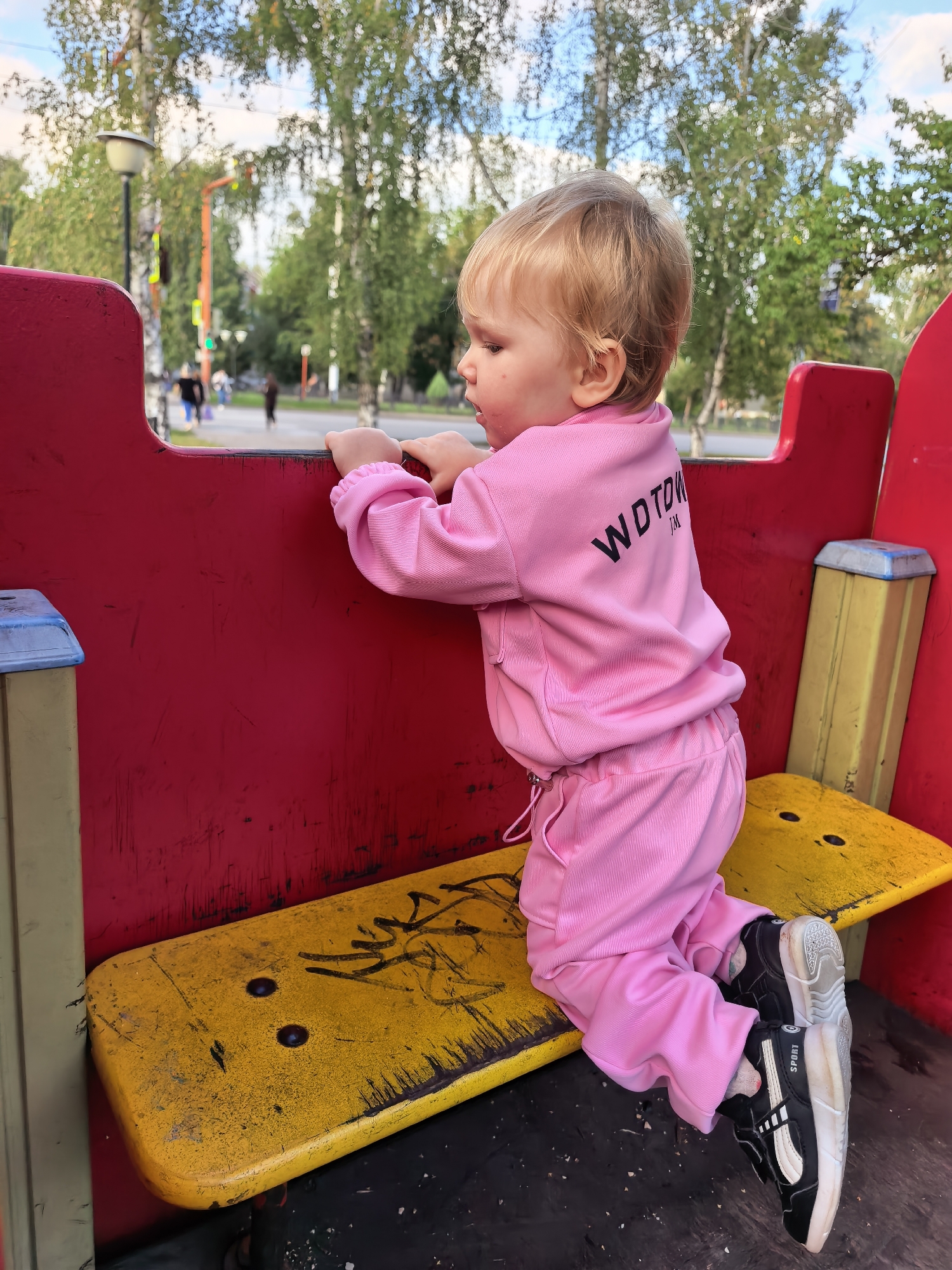 A young child with blonde hair, wearing a pink tracksuit, kneels on a yellow bench in an outdoor playground. The child leans forward, resting their hands on a red surface that appears to be part of a playground structure, possibly a train or similar vehicle. The child's face is turned to the side, looking down. In the background, there are trees, a lamppost, and blurred figures of people walking, suggesting an urban park or public space. The bench is weathered, with graffiti marks on its surface, and the ground is covered with dark material and scattered debris. The image is captured in a medium shot, focusing on the child and the immediate playground equipment. This scene is set in the city of Sterlitamak, Russia.