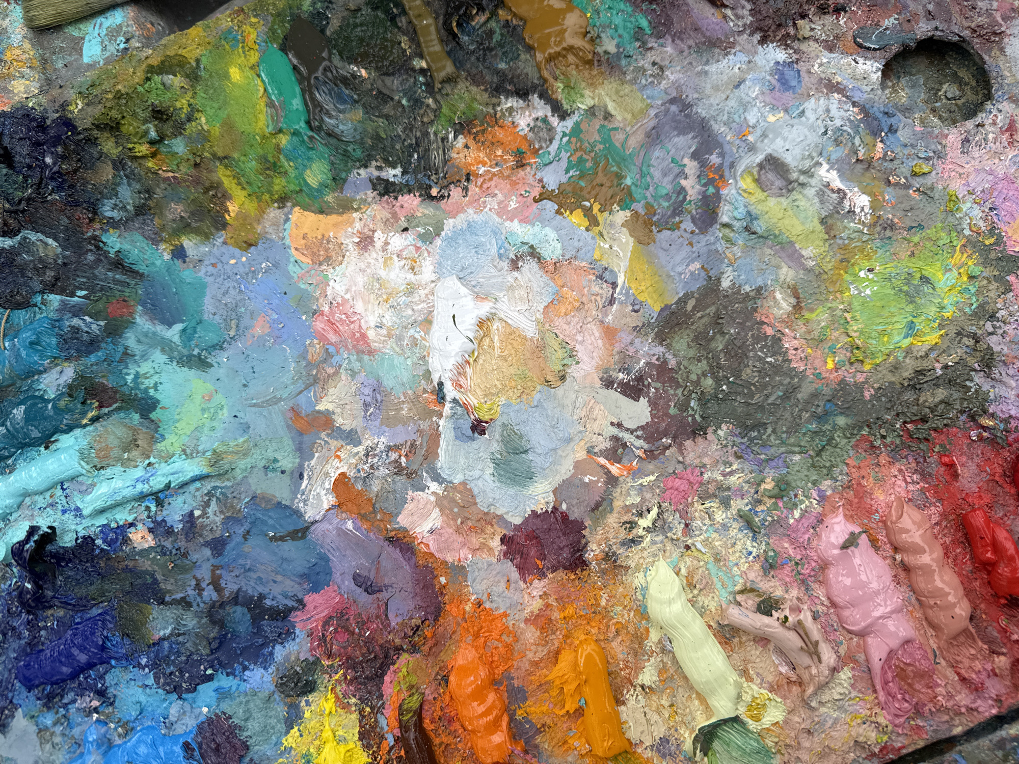 The image is a close-up, overhead view of an artist's palette, heavily laden with a variety of oil paints. The paints are textured and appear to have been mixed and applied with brushes, creating thick, impasto-like mounds and streaks.
The subjects are the oil paints themselves, which display a rich and diverse color palette. Hues range from deep blues, purples, and blacks to vibrant greens, yellows, oranges, pinks, and reds, with numerous shades of gray, beige, and white mixed in.
The activity taking place is implied to be the process of painting. The palette is a working surface, where colors are chosen, mixed, and prepared for application. The textures and layering of the paint suggest that this is not a new palette, but one that has been used for some time, with colors layered and blended from previous sessions.
Notable details include the thick, tactile quality of the paint, which suggests the use of oil paints and perhaps a style that emphasizes texture. The way the colors are spread and mixed indicates a dynamic and ongoing artistic process. There are no explicit location cues visible that would tie it to New Athos, Abkhazia, beyond the provided context. The emotional tone is one of creativity and artistic endeavor, with the vibrant colors suggesting a lively or expressive artistic practice.
There is no visible text in the image.