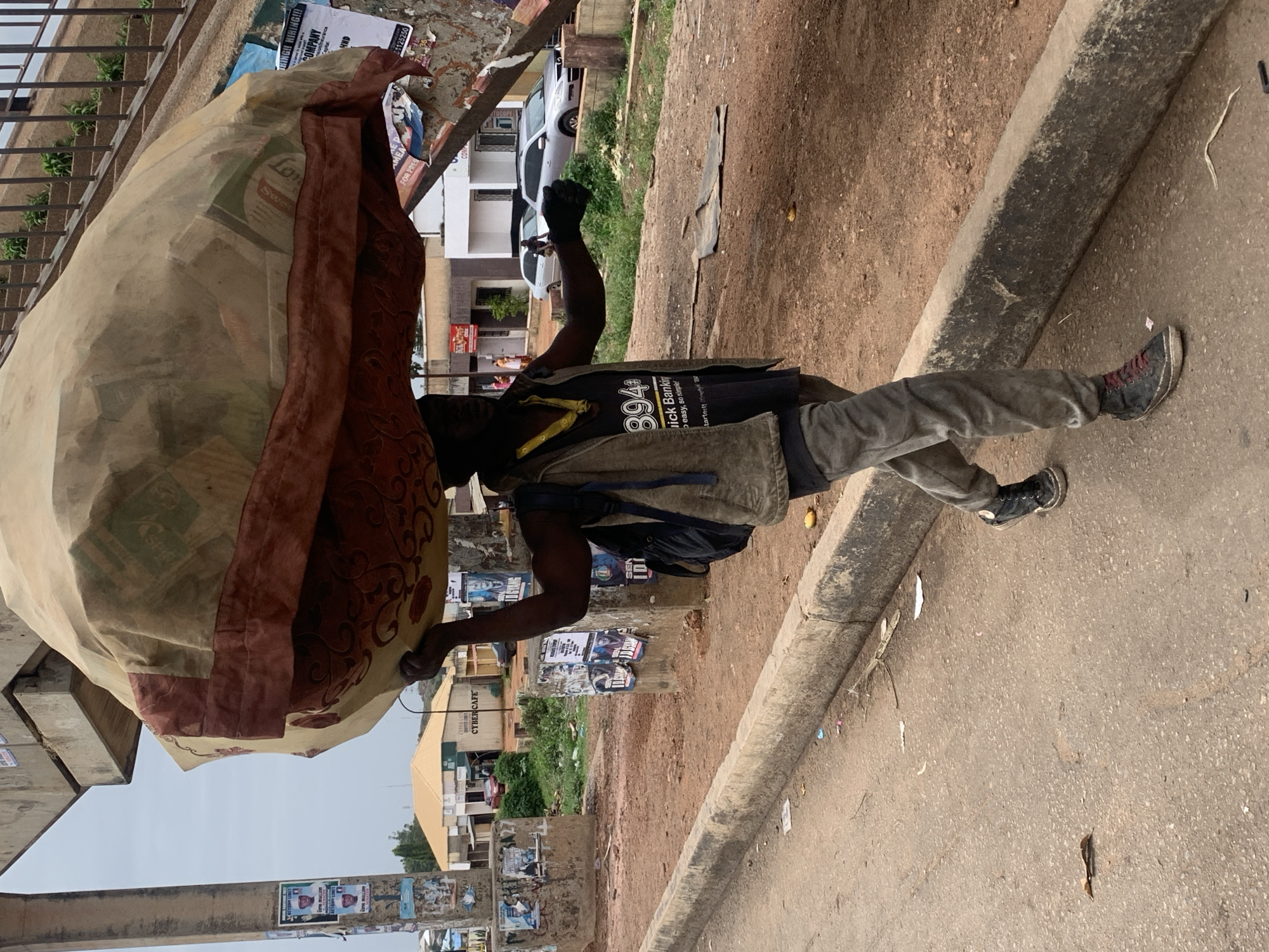 A man is walking along a street in Gura Top, Nigeria, carrying a large, bulging bag on his head. The bag is covered in a patterned cloth with a brown trim. He is wearing a dark t-shirt with a yellow lanyard, a vest, grey pants, and black sneakers with red laces. He has a backpack on his back. His right arm is raised in a fist.
The street is paved and has a grey concrete curb. There is trash and debris scattered on the road and along the curb. Buildings and trees are visible in the background, and posters are on a wall along the street. A Cyber Cafe sign is visible.