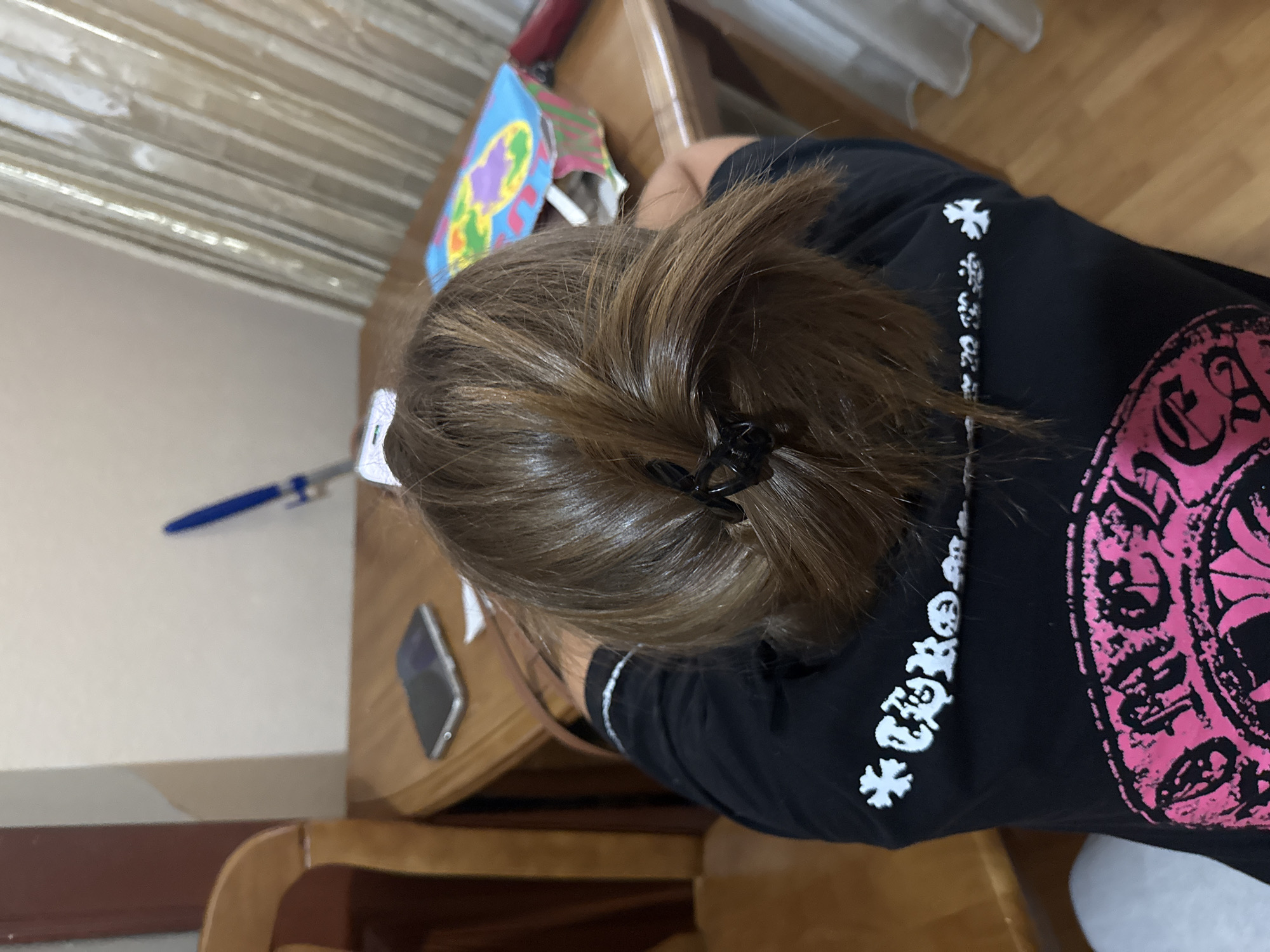 The image shows the back of a person's head with brown hair pulled back in a ponytail secured by a black clip. The person is wearing a black t-shirt with white and pink graphics. A wooden chair and a wooden table are visible. On the table, there is a smartphone, a blue pen, and a colorful box. The background shows a wall and curtains. The image was taken indoors, and there is no indication of the specific location in Dnipro, Ukraine.