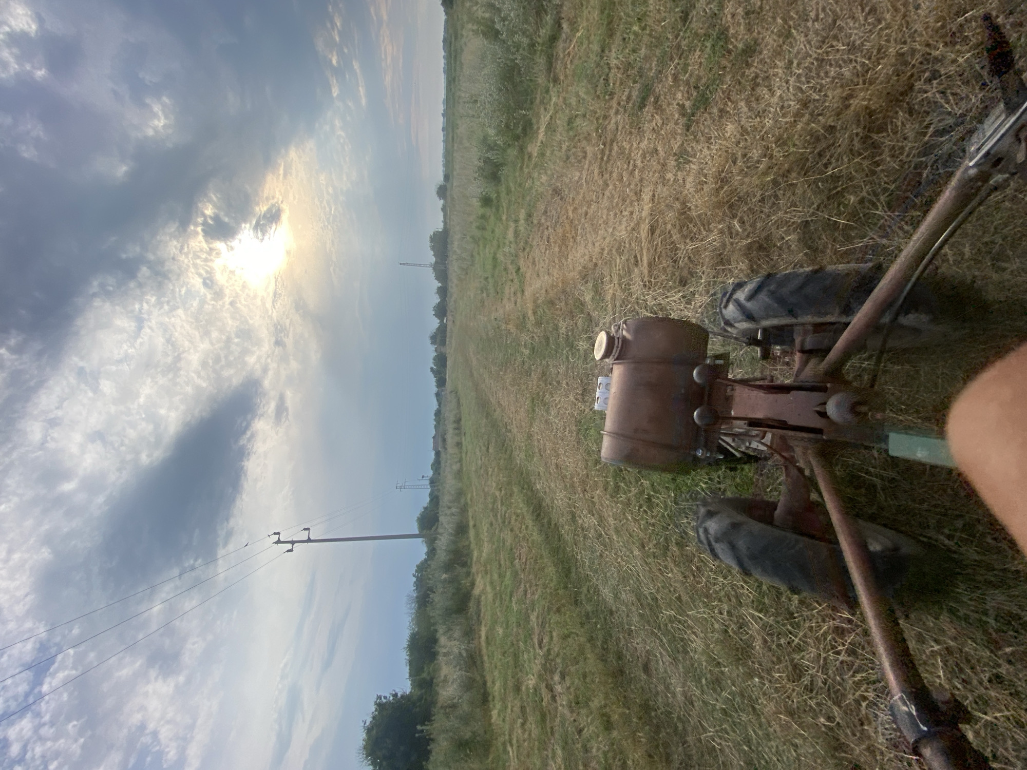 The image depicts a first-person perspective of someone operating a tiller in a field. The tiller is old and appears to be made of rusted metal, with two visible wheels and a fuel tank. The field is covered in dry, cut grass, suggesting recent agricultural activity. A dirt path leads away from the tiller into the distance. In the background, trees and vegetation line the sides of the field. Overhead, a cloudy sky with the sun peeking through dominates the upper portion of the frame. Power lines and utility poles are visible in the middle ground, extending towards the horizon. The scene appears to be set in a rural area, potentially on a farm or agricultural land in Brashlen, Bulgaria.