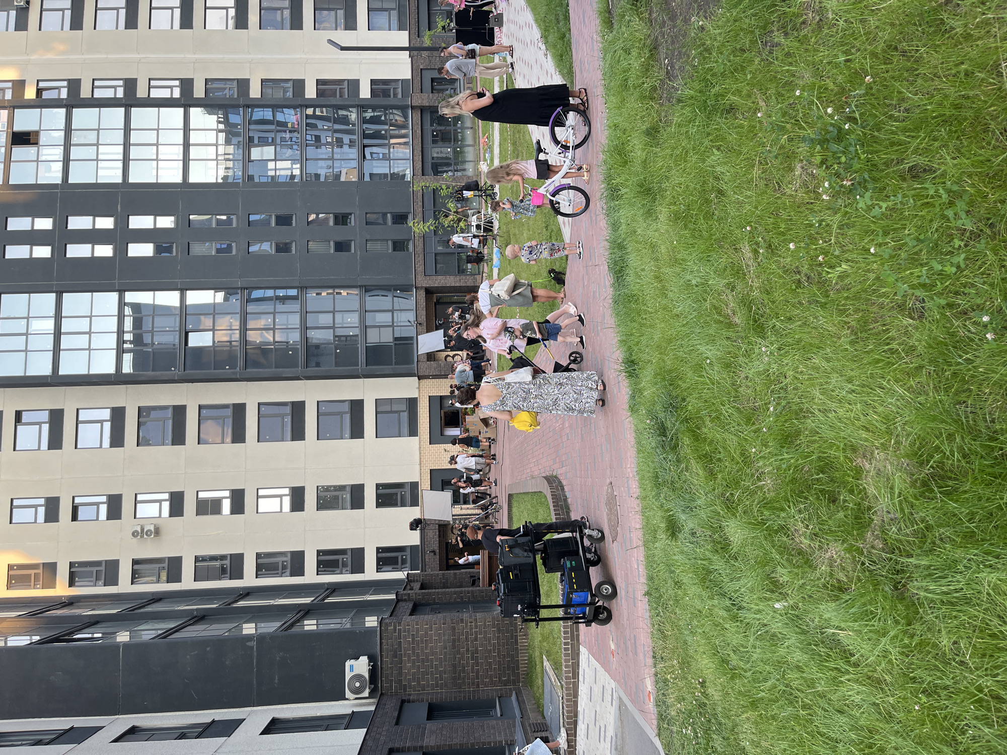 This is an outdoor, medium-shot, eye-level, full color, slightly hazy photograph of a courtyard in front of an apartment building. In the foreground, a man pushes a multi-wheeled cart laden with black equipment cases. The cart is on a brick pathway that runs through a grassy area. Several people are gathered in the middle ground, some of whom appear to be involved in a film or video shoot. There are lights and reflectors set up. Children are present, including two girls near a white bicycle. A woman in a black dress stands to the right of the frame, facing away from the camera. In the background, a large, modern apartment building with many windows dominates the scene. The overall impression is a busy, organized outdoor event.