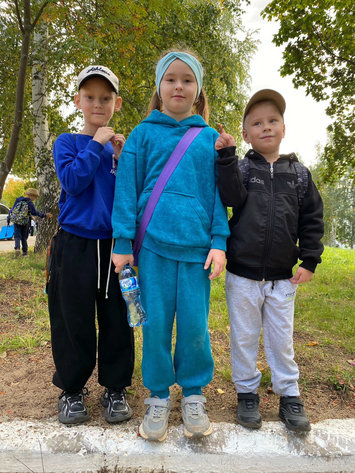 Three children are standing outdoors on a paved surface next to a grassy area with trees in the background. The child on the left, a boy wearing a blue sweatshirt and black pants, has a white baseball cap. The child in the middle, appearing to be a girl, is wearing a teal blue hooded sweatshirt and matching pants, with a purple strap across her chest and holding a bottle of water. The child on the right, a boy wearing a black jacket and gray sweatpants, has a beige baseball cap and a backpack. In the background, another child wearing a hat and backpack is visible near a vehicle. The foliage suggests an autumn setting. The image was taken in Noviy, Russia.