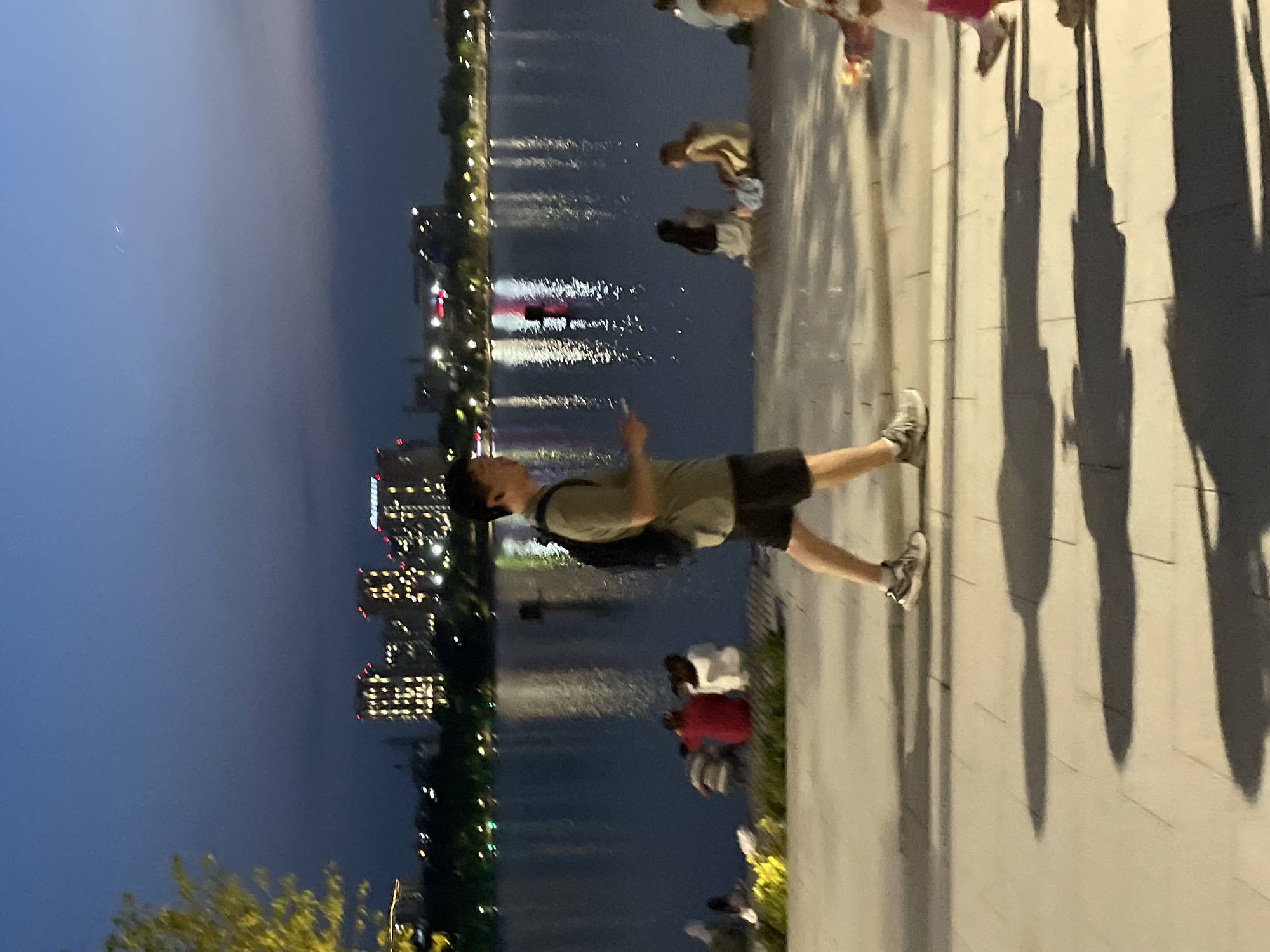 A man with a backpack walks on a paved path alongside a body of water at dusk. Buildings with illuminated windows are visible across the water, and their reflections shimmer on the surface. Several people are seated or standing near the water's edge, some appearing to be children. The sky is a deep blue, transitioning from the twilight hours. The scene is set in a city park or waterfront area, suggesting a public recreational space. While the image does not specifically indicate Almaty, Kazakhstan, it depicts a common urban landscape that could be found in many cities.