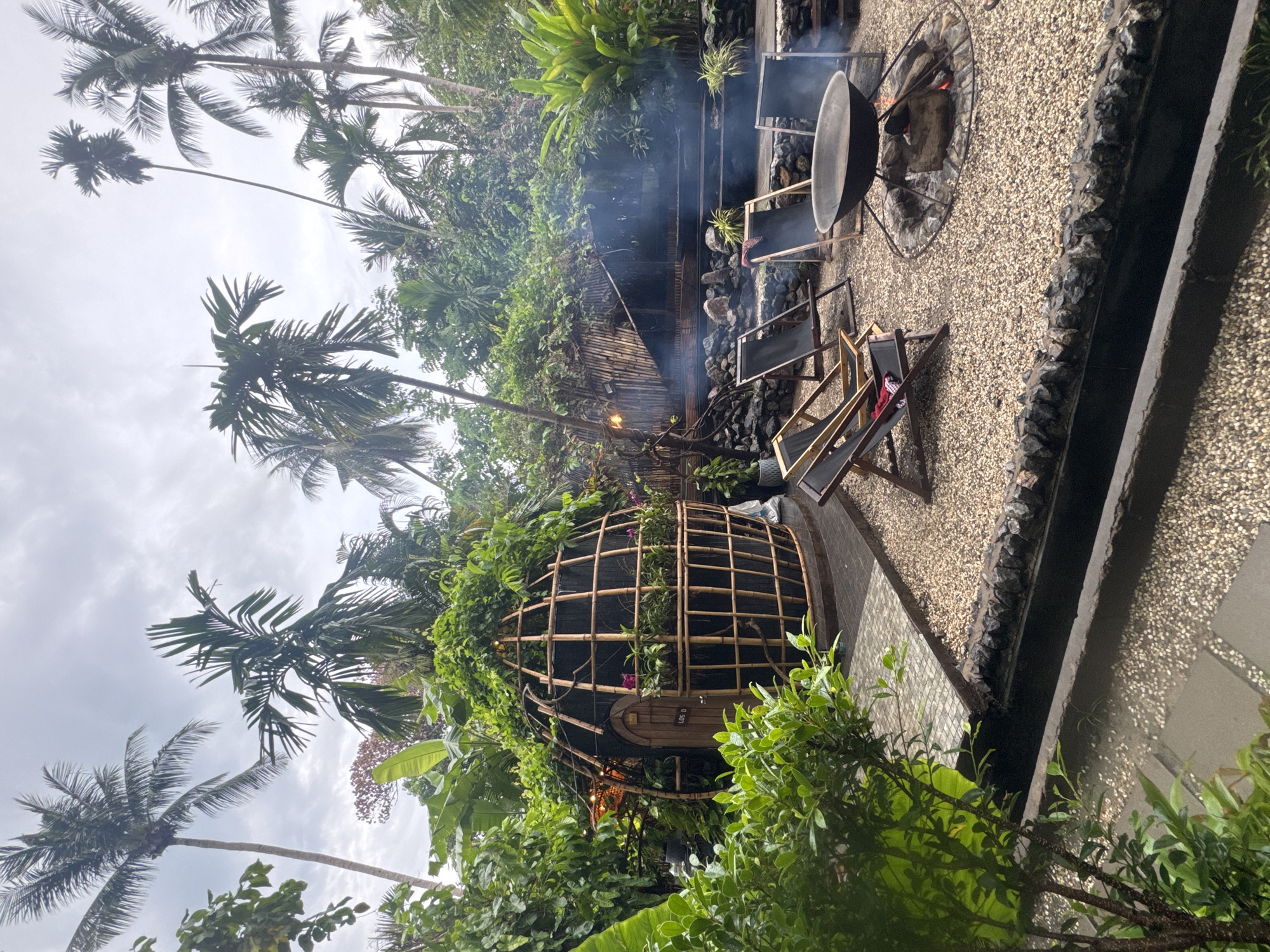 This photograph depicts a scene in what appears to be a landscaped outdoor space. The scene includes several distinct elements:
* **Vegetation:** The image is filled with lush greenery, including palm trees extending towards the sky and various other plants and bushes at ground level.
* **Domed Structure:** A unique, dome-shaped structure made of bamboo-like material is partially covered in climbing plants. It seems to have a wooden door.
* **Seating Area:** There is a seating area comprised of multiple wooden-framed chairs placed on a gravel surface.
* **Fire Pit:** A fire pit with a metal ring around it contains burning wood, producing smoke.
* **Gravel Area:** The seating and fire pit are situated on a gravel surface.
* **Border:** The gravel surface is bordered by a dark stone or concrete edge.
* **Sky:** The sky is visible through the trees and appears overcast.
The location is identified as Baan Sri Thanu, Thailand. While the image provides a clear view of the physical environment and objects within it, there are no visible people.