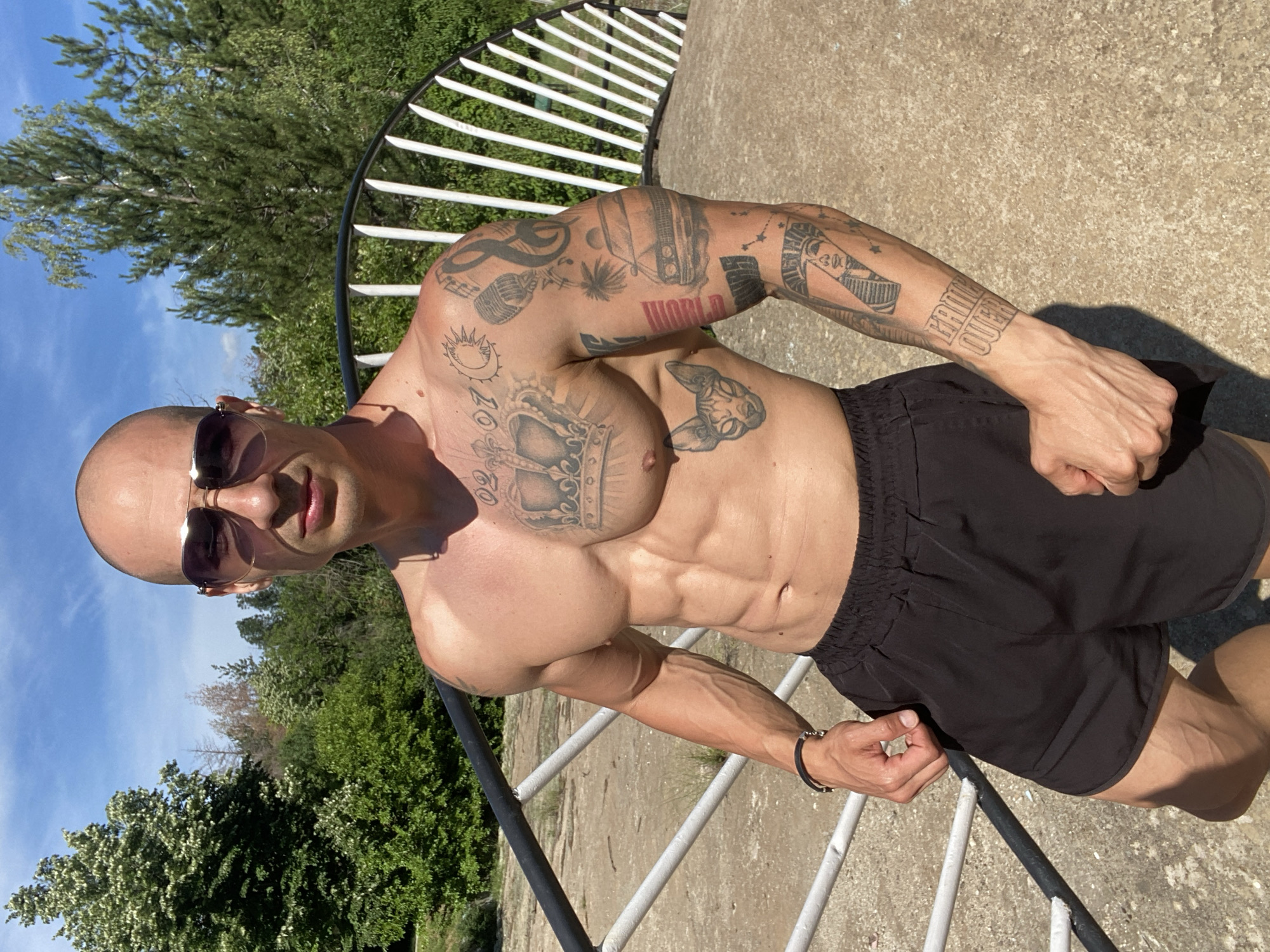 A shirtless, muscular bald man with sunglasses stands on a concrete path with a white railing in the background. He is wearing black shorts. He has tattoos on his chest and arms. The background shows lush green trees and a bright blue sky. The location is Lovech, Bulgaria.