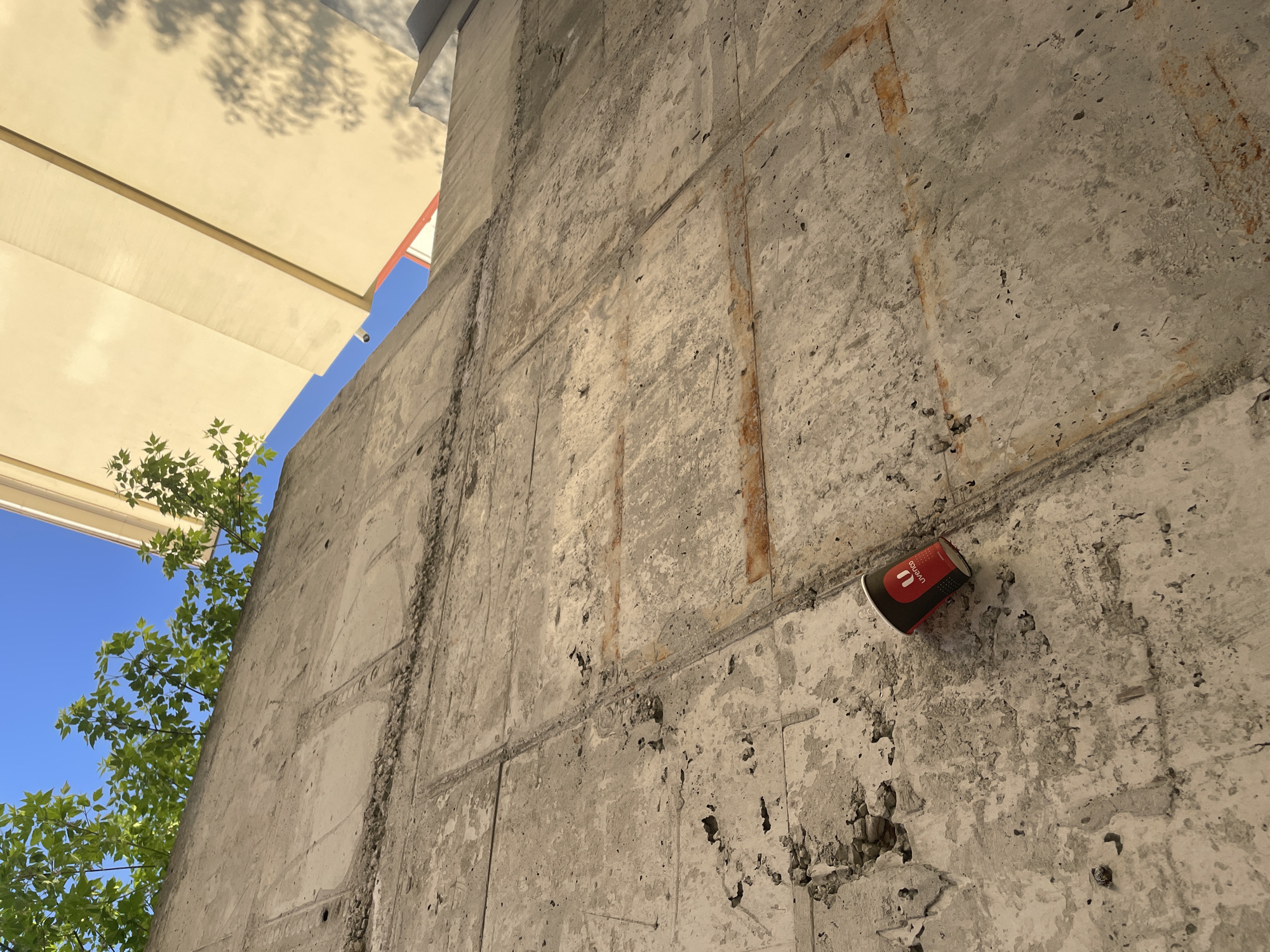 The image depicts an exterior concrete wall under an awning in Yekaterinburg, Russia. The wall has multiple vertical and horizontal indentations, indicating previous construction seams or repairs. A discarded red and black "Uralsky Uvelirny Zavod" (Uvelirco) paper cup is affixed to the wall, at approximately eye level, suggesting littering. Above the wall, branches of a tree are visible against a blue sky. The awning casts a shadow on the upper left of the image. There are no people visible in the image.