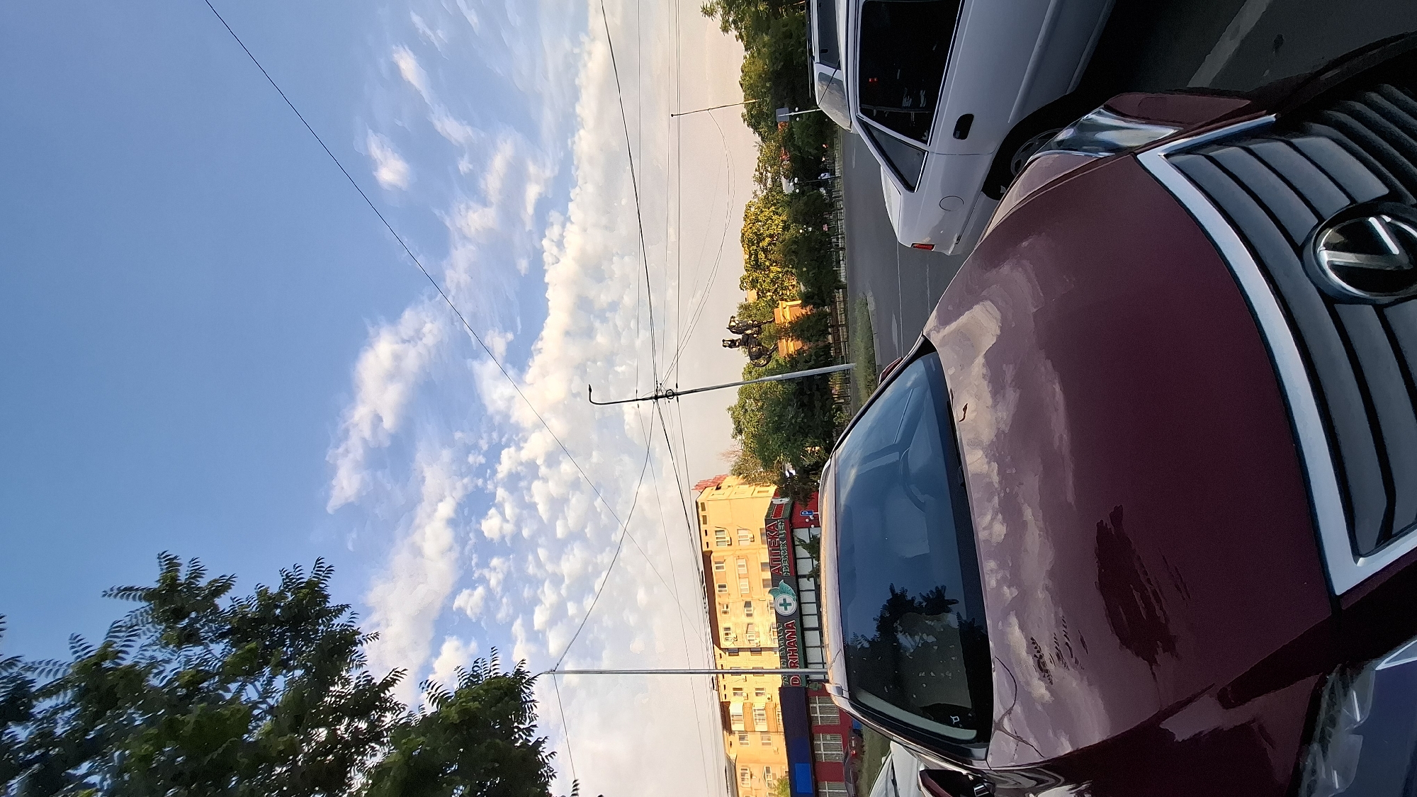 A low-angle shot captures a street scene in Shymkent, Kazakhstan, oriented vertically. The foreground is dominated by the dark red hood and windshield of a car, reflecting the sky and surrounding environment. In the middle ground, a white car is parked, and further back, a multi-story yellow building with visible windows and balconies stands. A sign on the building reads "APTEKA DORIHANA" with a green cross, indicating a pharmacy. Trees line the street and form a backdrop to the building. The sky is a vibrant blue with scattered white clouds, suggesting fair weather. A power line runs diagonally across the upper portion of the image. The lighting suggests it is either early morning or late afternoon. The overall scene depicts a typical urban street in Kazakhstan.