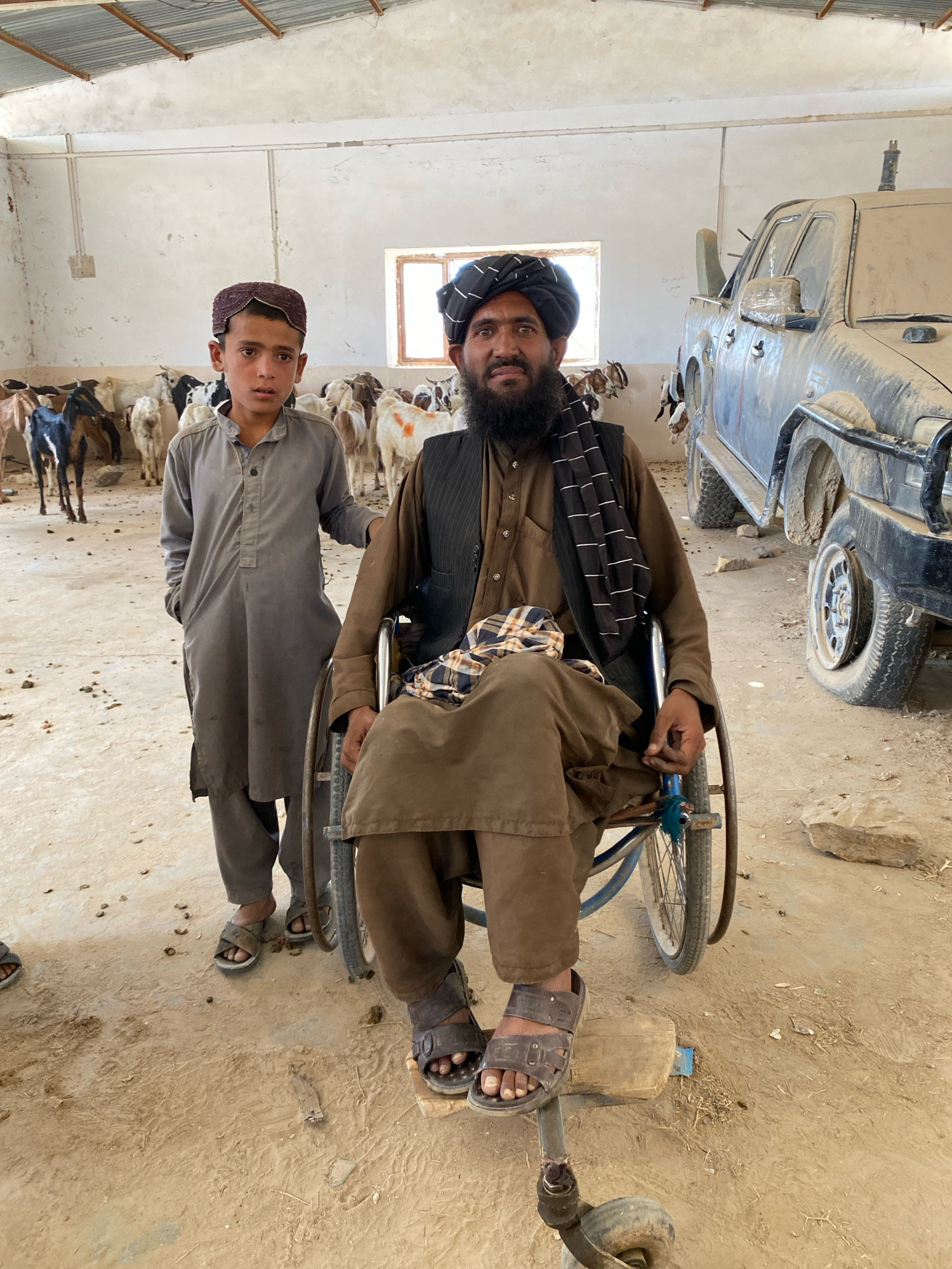 In the city of Pasab, Afghanistan, a young boy stands to the left of a man seated in a wheelchair. Both are dressed in traditional clothing, with the boy wearing a grey tunic and pants and a small embroidered cap. The man is in a brown tunic, vest, and turban, with a checkered cloth draped over his legs. Behind them, numerous goats of various colors are visible, suggesting an animal enclosure or farm. A dirt floor covers the ground, and a window is present in the back wall, along with a portion of a dusty, dark-colored vehicle. The environment appears to be a somewhat rudimentary indoor space with a metal roof. The overall scene conveys a sense of everyday life in a rural or semi-rural Afghan setting.
