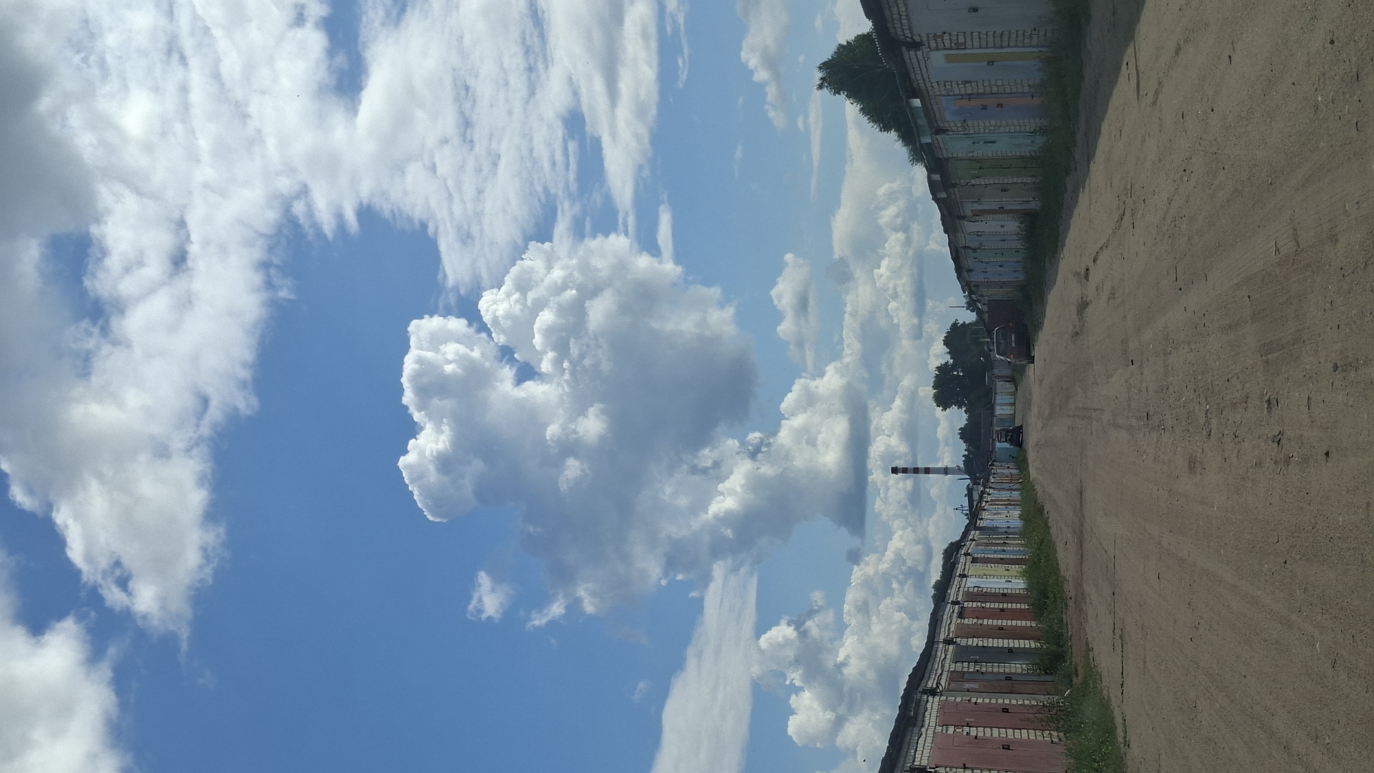The image depicts a row of garages on a dirt road in Maladzyechna, Belarus. The garages are constructed of brick and have various colored doors. The road is unpaved and appears to be well-traveled. In the distance, a tall, red and white striped chimney rises above the garages, suggesting the presence of an industrial facility. The sky is blue with scattered white clouds, some of which are large and puffy. Two dark-colored cars are parked on the road between the garages. The overall scene is one of urban infrastructure in a relatively undeveloped area.
