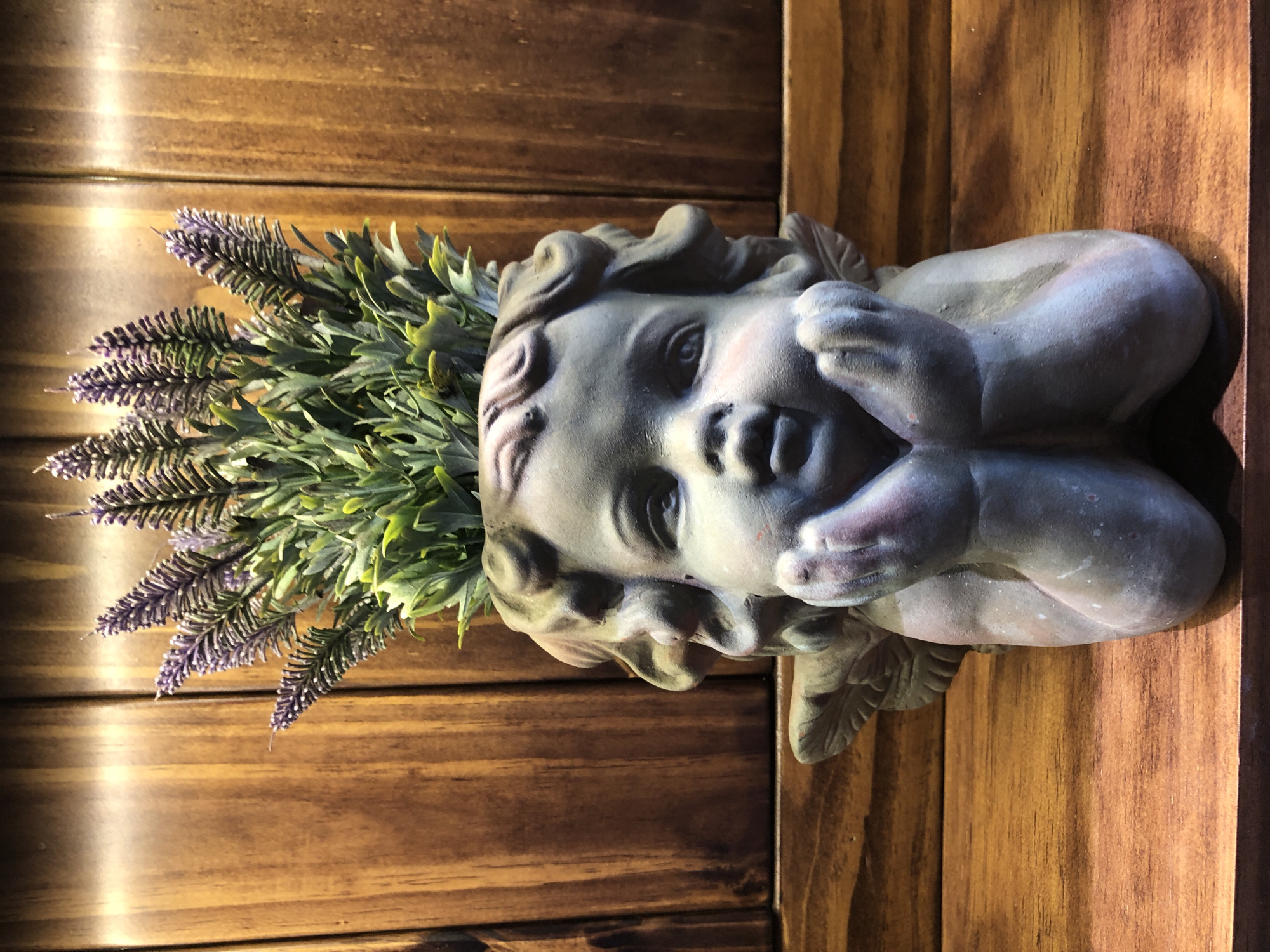 The image shows a close-up of a decorative pot shaped like a cherub with a bouquet of artificial lavender plants growing from its head. The pot is made of a gray, weathered material and is positioned on a wooden surface. Behind the pot is a wooden background with vertical planks. The lighting suggests an indoor setting, possibly a shelf or display area. There are no people or any indication of a specific location in Đà Nẵng, Vietnam.