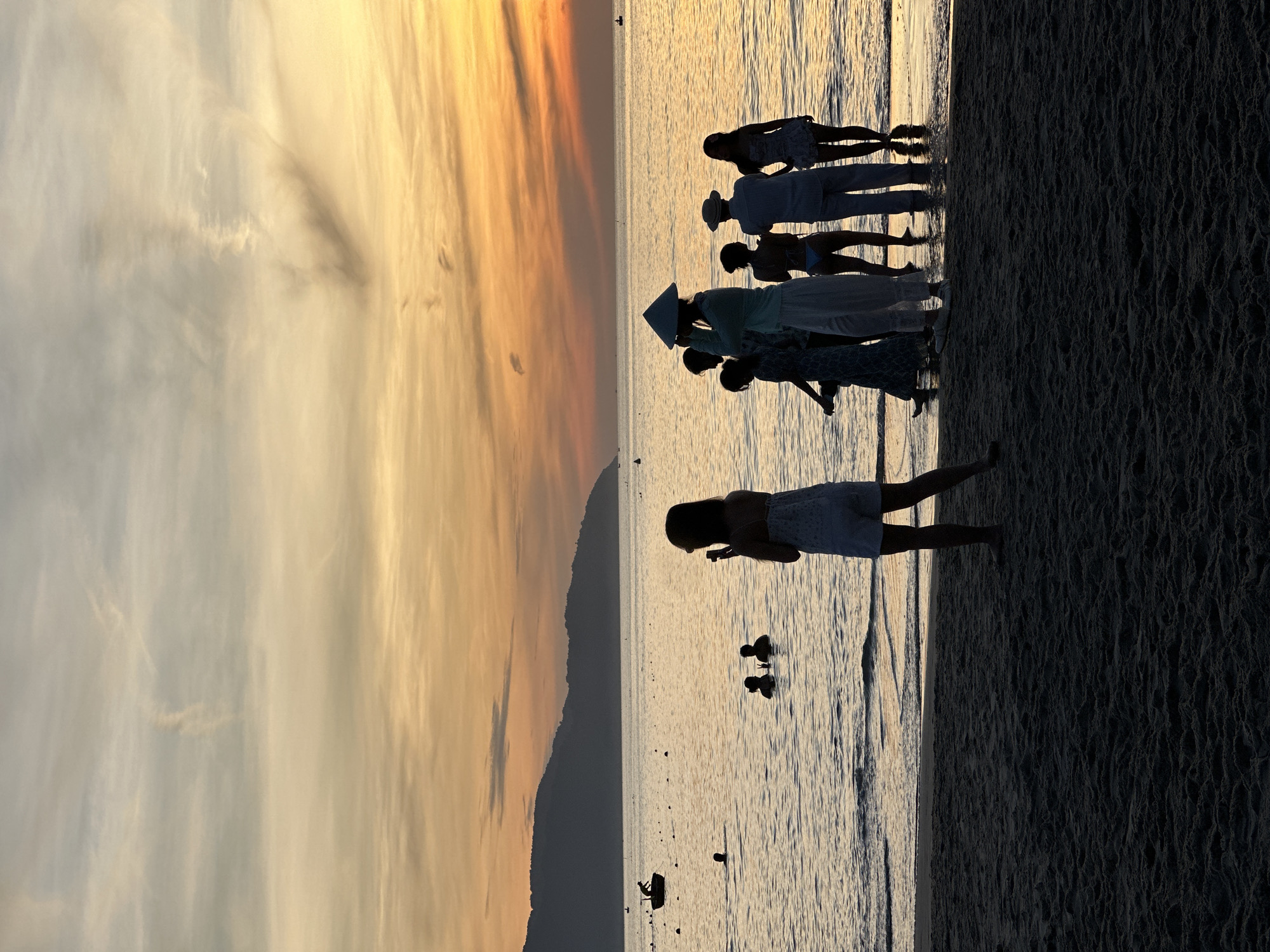 At sunset on a beach in Da Nang, Vietnam, silhouettes of people gather near the water's edge. A group of seven individuals stand in the shallow sea, some facing the ocean and others looking at the shore. One person in the foreground, facing away from the camera, holds up a phone, possibly taking a photo. A lone individual stands on the wet sand, also facing the water. Further out, two people are partially submerged in the ocean, and a small boat with a person in it is visible in the distance. The sky is painted with warm hues of orange and yellow, characteristic of a sunset, with mountains forming a dark silhouette on the horizon. The sand in the foreground is dark and textured, indicating it is wet from the tide.