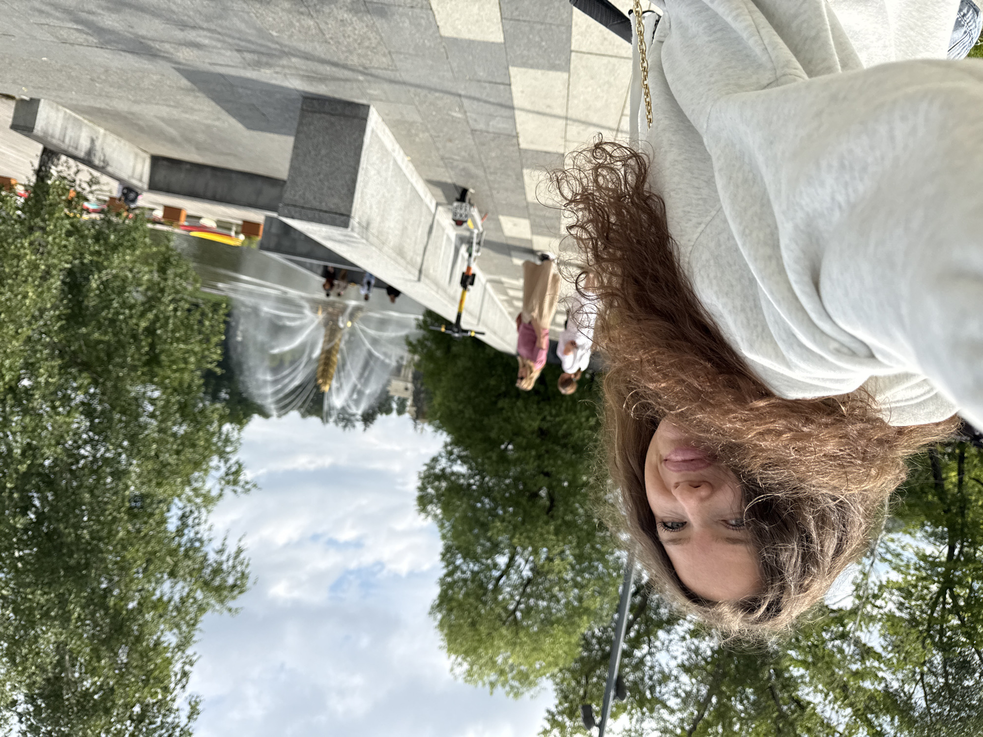 A woman with long, curly brown hair takes a selfie outdoors. She is wearing a light-colored hooded sweatshirt. In the background, a large fountain sprays water high into the air, with trees and a cloudy sky visible behind it. A few other people are walking or sitting near the fountain. An electric scooter is parked on the walkway. The scene appears to be in a park or public outdoor space, possibly in Moscow, Russia, given the architecture of the fountain which resembles the Friendship of Nations fountain in VDNKh Park.