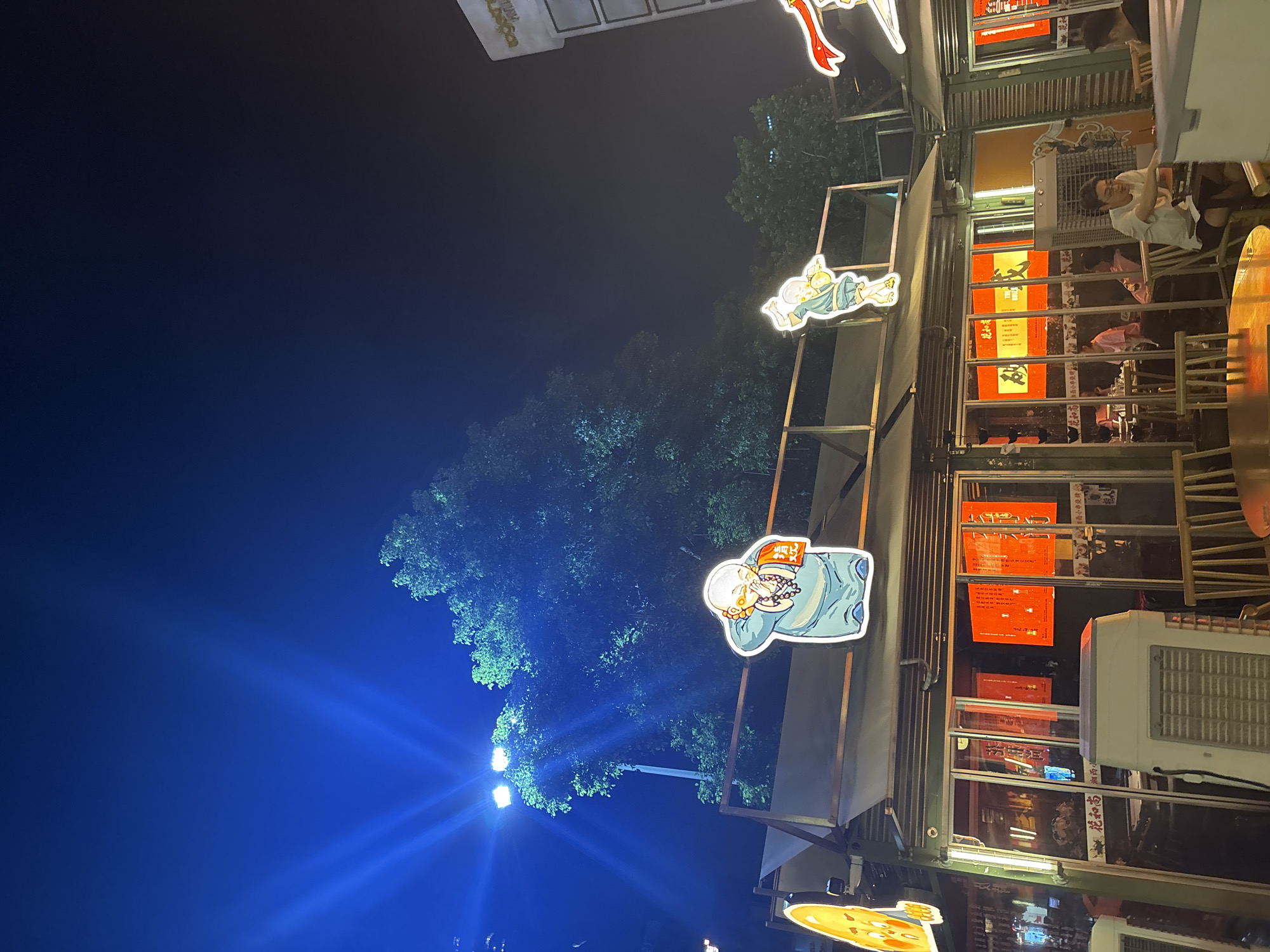 This is a nighttime, outdoor shot of a street scene, possibly a restaurant or shop in Haizhu District, China. The image is vertically oriented and has a dark blue, starry sky with bright blue spotlights illuminating some trees on the left side. On the right, a building with multiple windows is visible. The building has a yellowish-orange facade and is adorned with illuminated decorative cutouts of cartoonish figures, one appearing to be a monk. Some of the cutouts are attached to a metal frame structure. Through the windows, it appears to be an interior space with tables and people, suggesting a dining establishment. Several red signs with Chinese characters are visible behind the glass. One cutout, of a monk figure, is prominently featured on the left side of the frame. The overall mood is lively and festive due to the colorful decorations and lights, despite the darkness of the night. The presence of streetlights and illuminated signs suggests it is a commercial area.