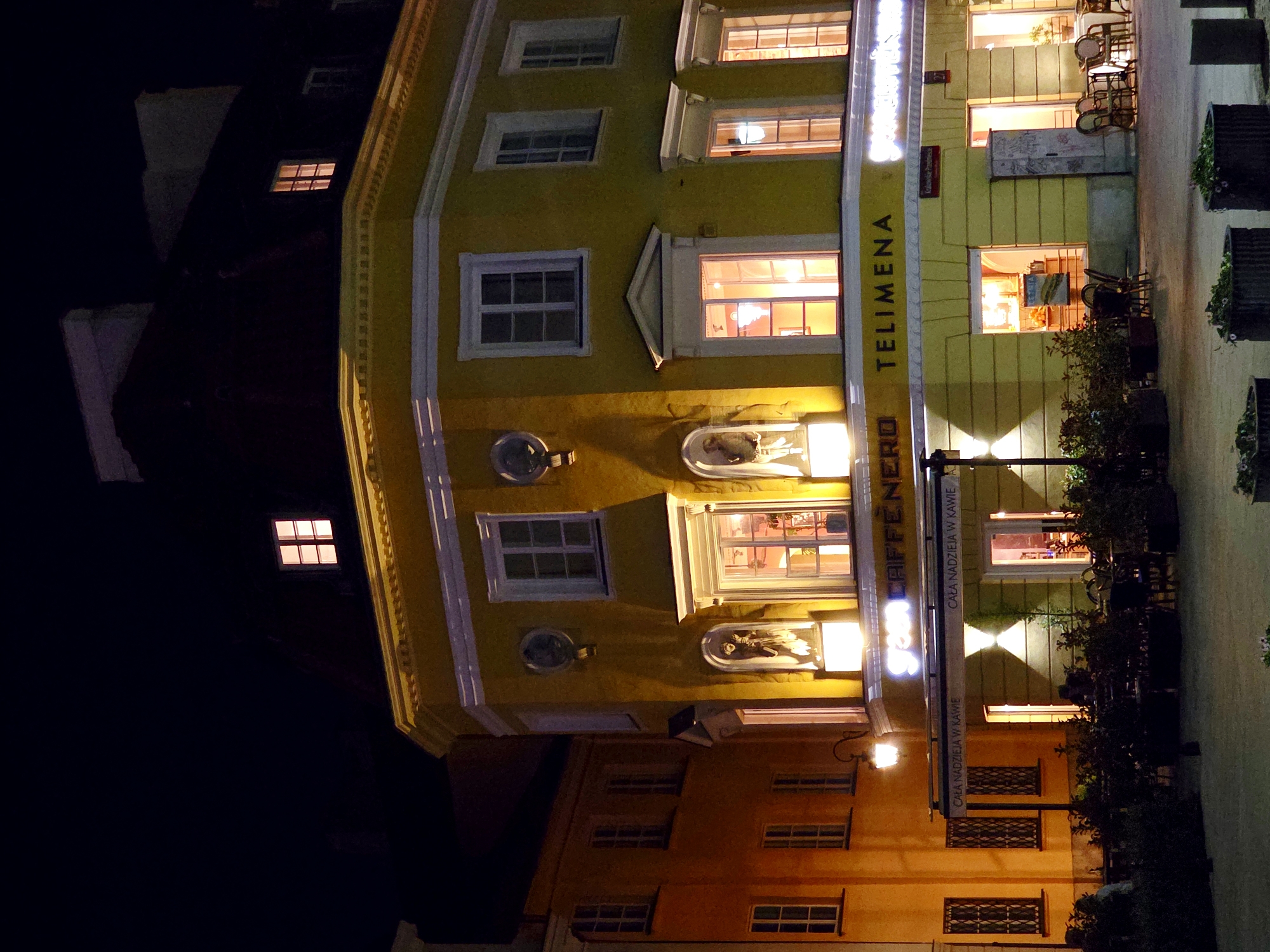 This is a nighttime photograph of the historic Old Town of Warsaw, Poland, featuring a bright yellow building with multiple windows illuminated from within. The building has a red-tiled mansard roof, characteristic of Warsaw's architecture. A prominent sign on the building reads "green CAFFÈNERO" and "TELIMENA," indicating a cafe or restaurant. The street level displays an outdoor seating area with tables and chairs, surrounded by dark green foliage in planters. The surrounding buildings and streetlights cast a warm glow on the scene, creating a cozy and inviting atmosphere. The image captures the charm of Warsaw's historic district at night, with a focus on the building's architectural details and the lively ambiance of the cafe.