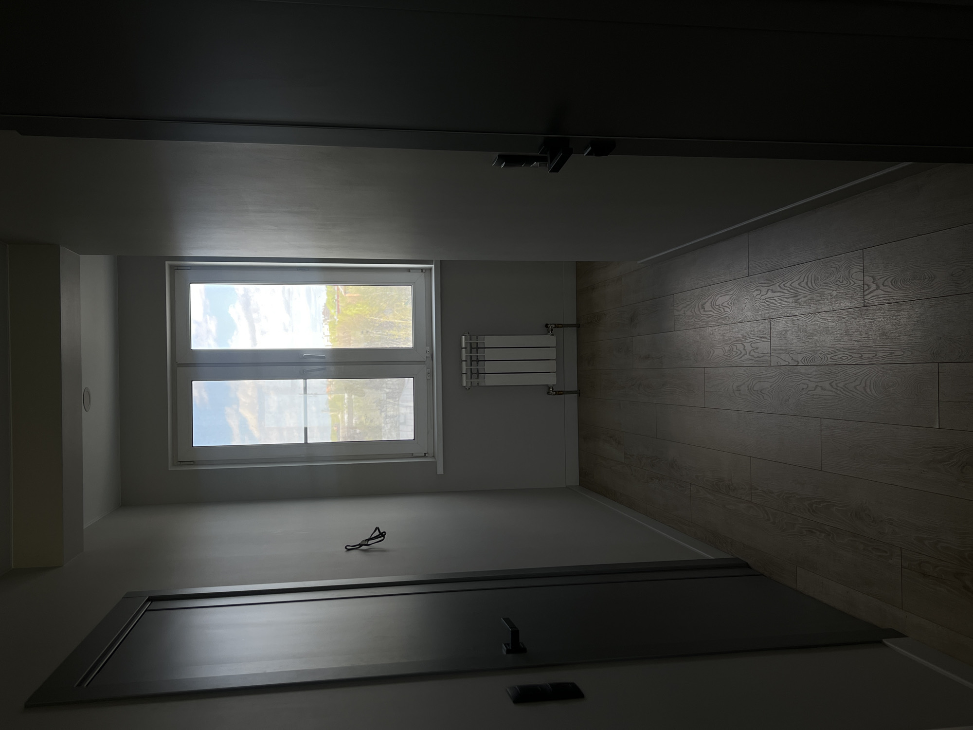 The image depicts the interior of a room, potentially in Vologda, Russia (no visual confirmation available), focusing on architectural details.
**Location:** The interior of an empty room.
**Environment:** The room has light-colored walls and what appears to be a light-colored wood-patterned floor. There is a window with two panes, through which an exterior scene is visible (trees and sky). A radiator is mounted on the wall near the window. The room appears to be illuminated by natural light coming through the window.
**Objects:**
* Window with two panes.
* Radiator on the wall.
* An object, possibly glasses, is lying on the floor in the foreground.
* Door(s) are partially visible on both top and bottom framing the view.
**People:** No people are visible.
**Actions/Interactions:** There is no indication of ongoing action or interaction.
**Overall Impression:** The scene is a neutral, empty room with a focus on architectural elements.