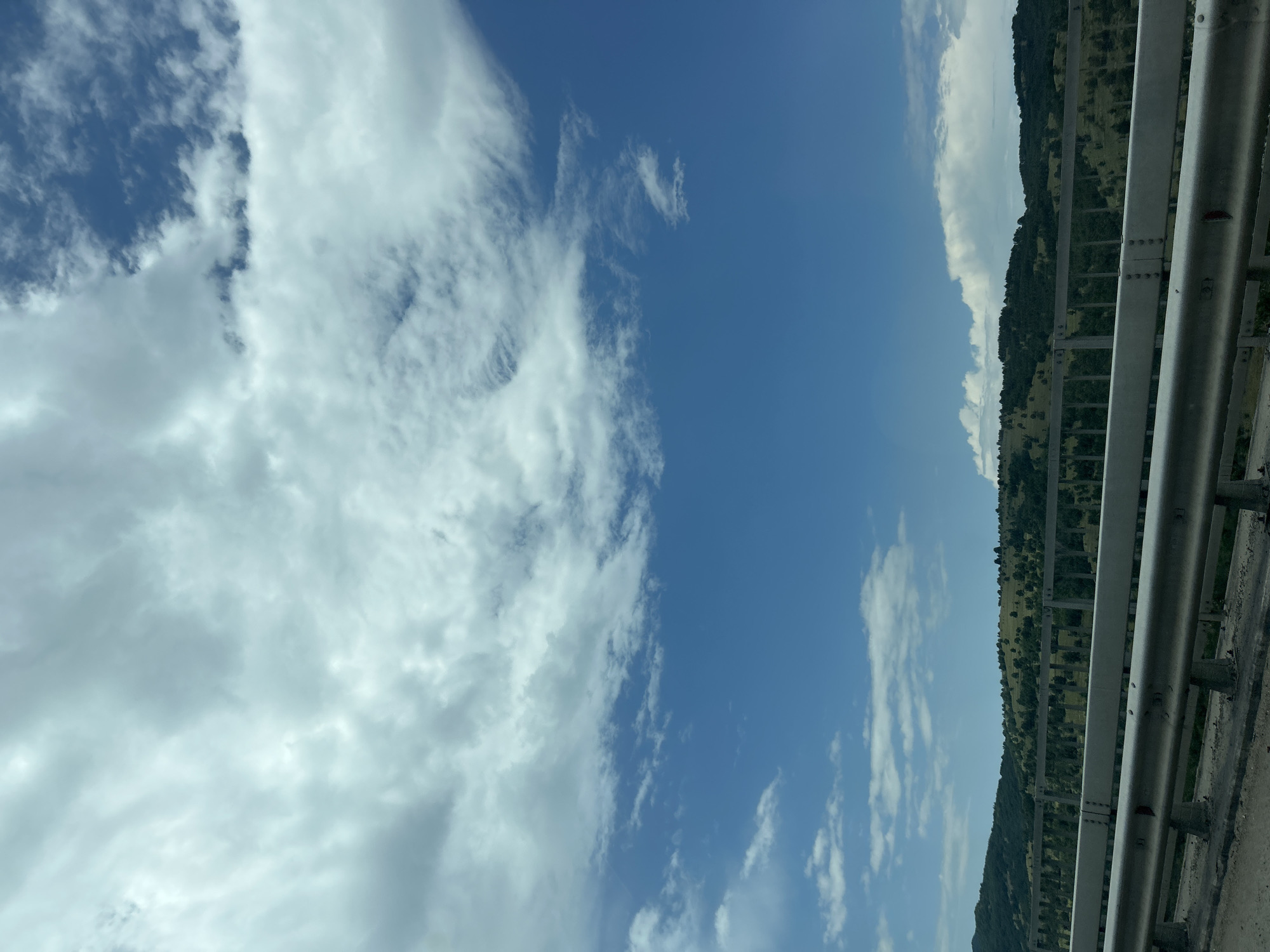 This image captures a wide-angle view of a natural landscape and human-made infrastructure in Gorno Varshilo, Bulgaria. The upper two-thirds of the frame are dominated by a dynamic sky, featuring a blend of dense, white-to-grey stratocumulus clouds in the upper left transitioning to patches of clear, deep blue sky and scattered lighter cumulus and cirrus clouds towards the right and middle. The horizon line is visible in the lower third, revealing rolling hills covered with varied vegetation. These hills are predominantly green, indicating lush growth, with a mix of grassy areas and numerous scattered trees or small clusters of woodland. In the immediate foreground, a substantial metallic road barrier or bridge railing spans the width of the frame. This barrier consists of multiple horizontal silver-colored rails secured by visible bolts and supported by vertical posts, anchored into a concrete structure. The concrete base shows signs of wear and minor debris. No people, animals, or vehicles are present in the scene.