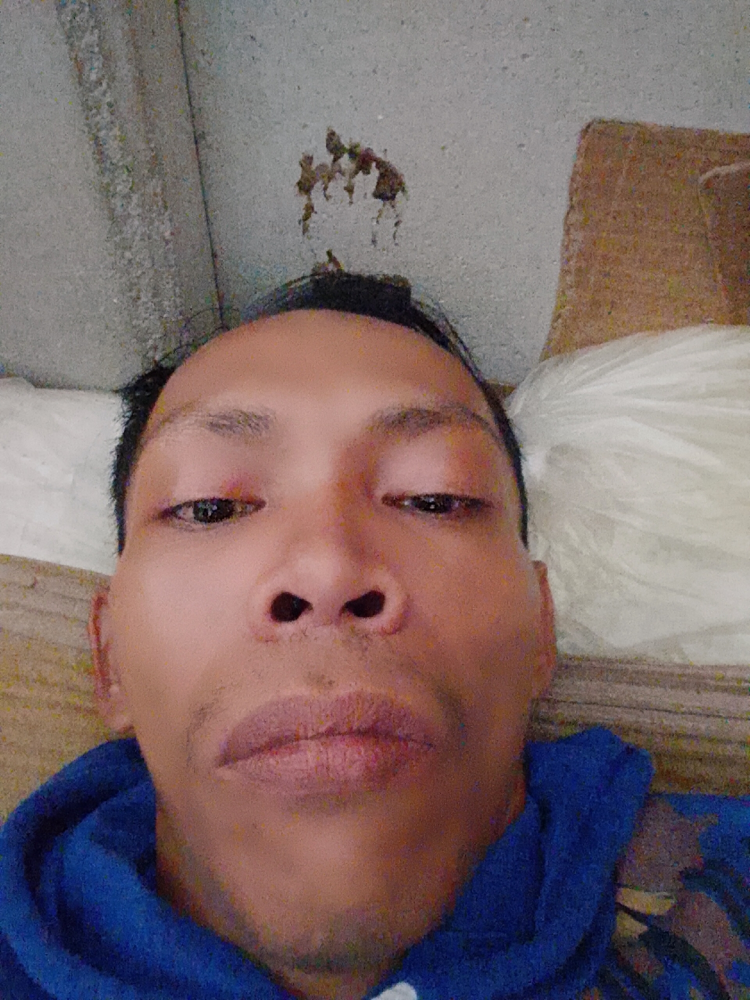 A selfie, taken from a low angle, of an individual with dark hair and brown eyes. The person's face is centered in the frame. They are wearing a blue hooded sweatshirt. The background includes a white pillow and a brown object; the ceiling is concrete and displays what appears to be dried organic material. The location is Bukit Ketok, Indonesia.