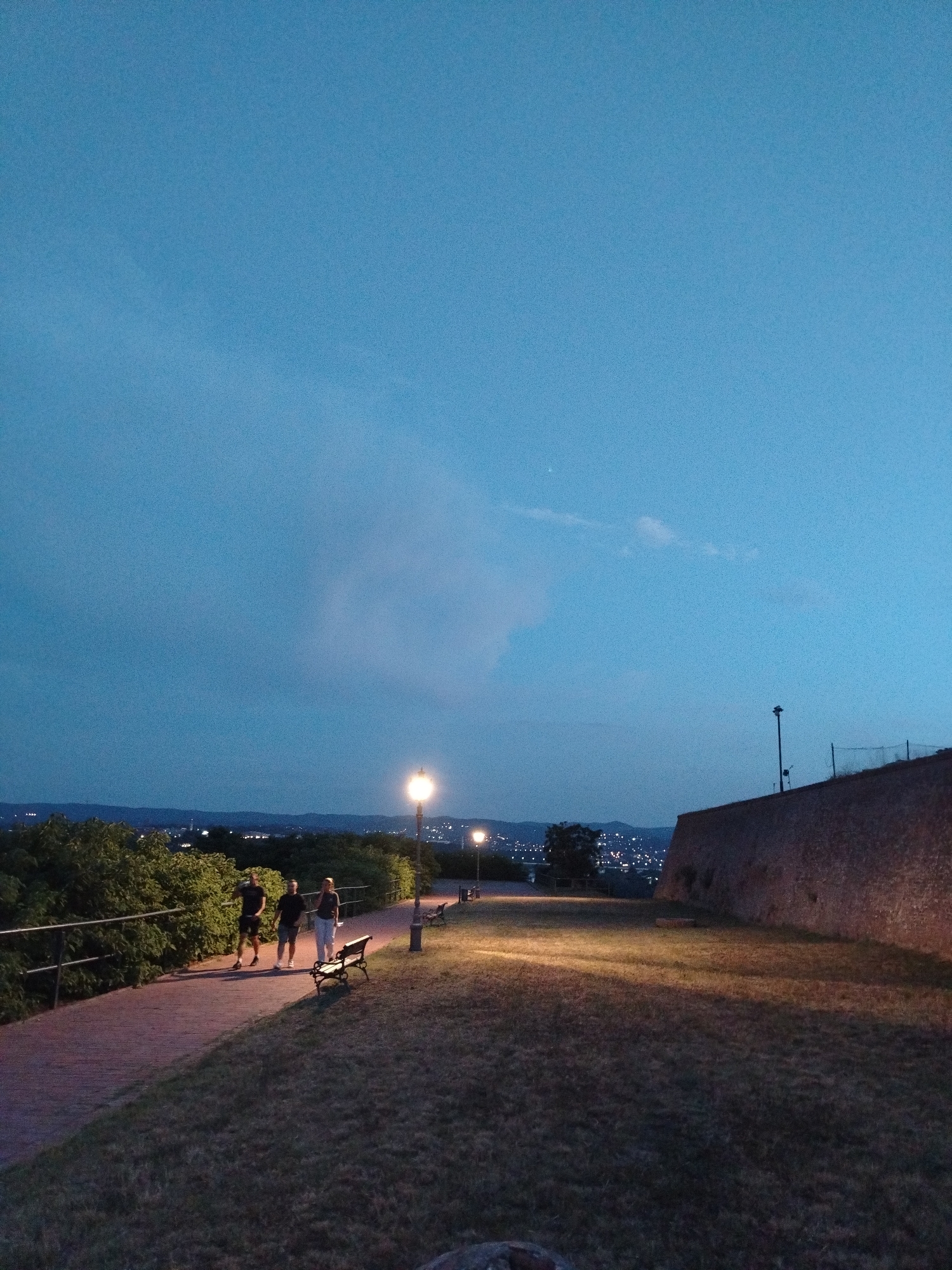 The image depicts an evening scene at Petrovaradin Fortress in Petrovaradin, Serbia. The sky is a deep blue, transitioning to a lighter shade towards the horizon, with a wispy cloud formation visible in the upper left. In the foreground, a grassy area with long shadows from streetlights is illuminated by warm light. A paved pathway runs diagonally across the scene, bordered by lush green bushes on the left. Three individuals are walking along the pathway. Two men, one in a dark t-shirt and shorts and the other in a black t-shirt and dark shorts, are walking side-by-side. A woman in a white top and light-colored pants walks slightly behind them. A park bench is positioned near the pathway. Further along the pathway, more benches and lampposts are visible. To the right of the pathway, a textured brick wall, characteristic of the fortress's architecture, rises. In the background, a city skyline with numerous lights twinkles against the darkening landscape, suggesting the presence of the city of Novi Sad across the Danube River. A tall, slender pole with a light fixture or camera is situated atop the wall on the right, and a fence is visible behind it. The overall atmosphere is serene and tranquil, capturing a moment of leisure as dusk settles over the historic fortress.