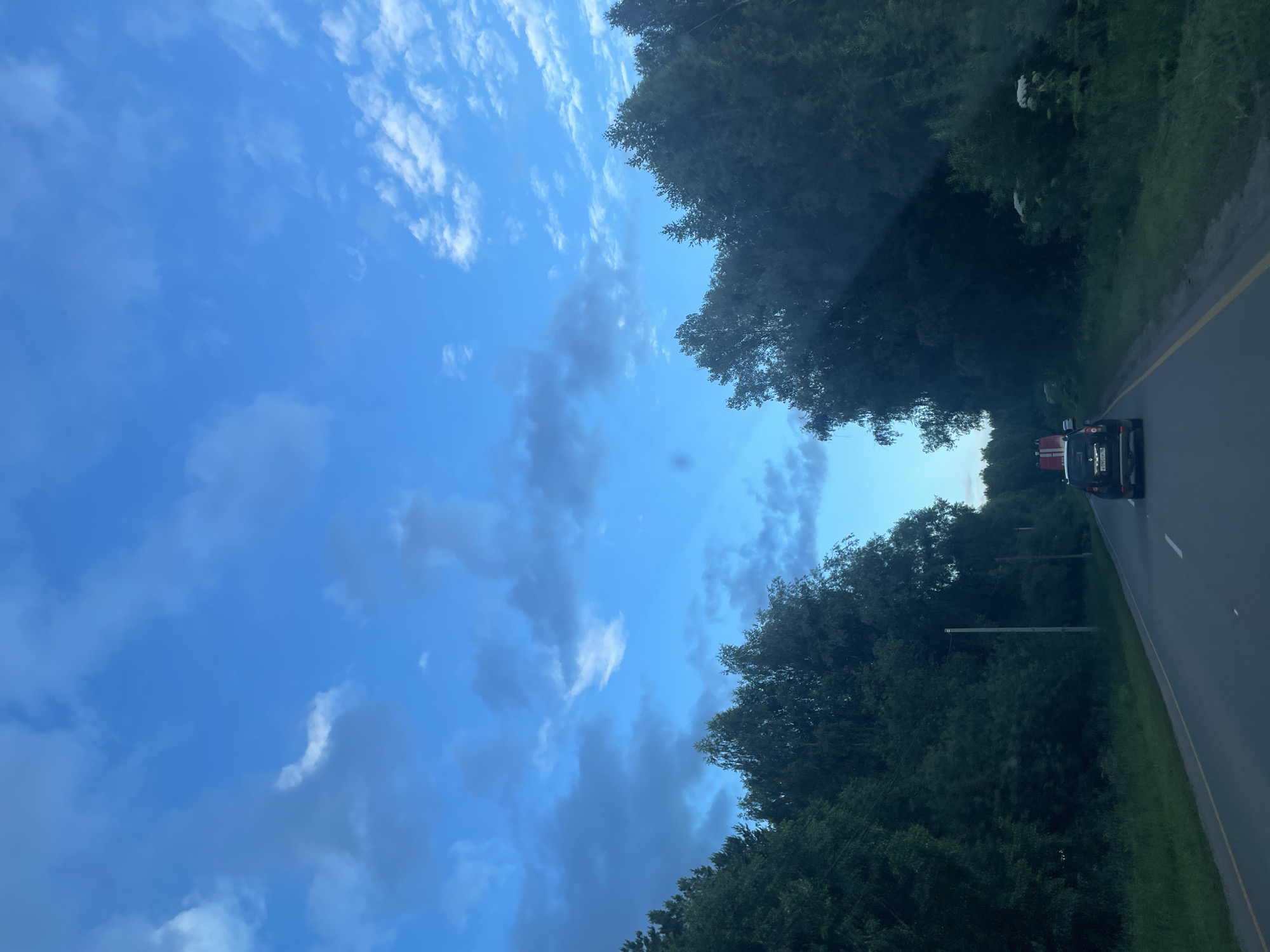 The media file depicts a view from inside a vehicle traveling along a road lined with dense trees. The sky is a vibrant blue, scattered with white and gray clouds, suggesting fair weather with some cloud cover. It appears to be daytime, likely late afternoon or early evening, given the soft lighting.
The primary focus is the road and its surroundings. On the right side of the frame, a dark-colored vehicle, possibly a truck or SUV with red markings on the back, is visible driving ahead. The road itself has a yellow line marking the edge and a white line indicating a lane. The trees on both sides of the road form a green canopy, creating a tunnel-like effect.
In the distance, on the right side of the road, there are some white objects in the trees, their nature is unclear. There are also utility poles visible along the roadside. The scene conveys a sense of travel through a natural, possibly rural or forested, area. The overall mood is serene and peaceful, capturing a moment of a road trip. No people are directly visible. The orientation of the image is landscape, with the road running from the bottom right towards the top left. There is some text visible on the license plate of the vehicle in front, but it is too blurry to read.
The location context provided is Камское, Russia.