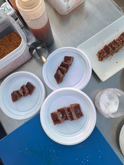 The image presents a flat lay scene, likely on a metallic food preparation surface, possibly in Barbate, Spain, given the food items. Three small white plastic plates each hold several pieces of what appears to be sliced, dark red tuna. The tuna is cut into roughly square pieces and arranged linearly on the plates. One plate has a light sprinkling of salt. To the left of the plates, there is a rectangular container filled with a dark granular substance. A squeeze bottle containing a reddish-brown liquid, likely a sauce, is positioned above the container. There is also a small metal scoop. To the upper right, there is a rectangular white plate with a strip of tuna coated with a mixture of what appears to be sesame seeds or similar small grains. Also, in the bottom right, there is a mound of salt in a clear plastic container. A blue surface is visible in the foreground.