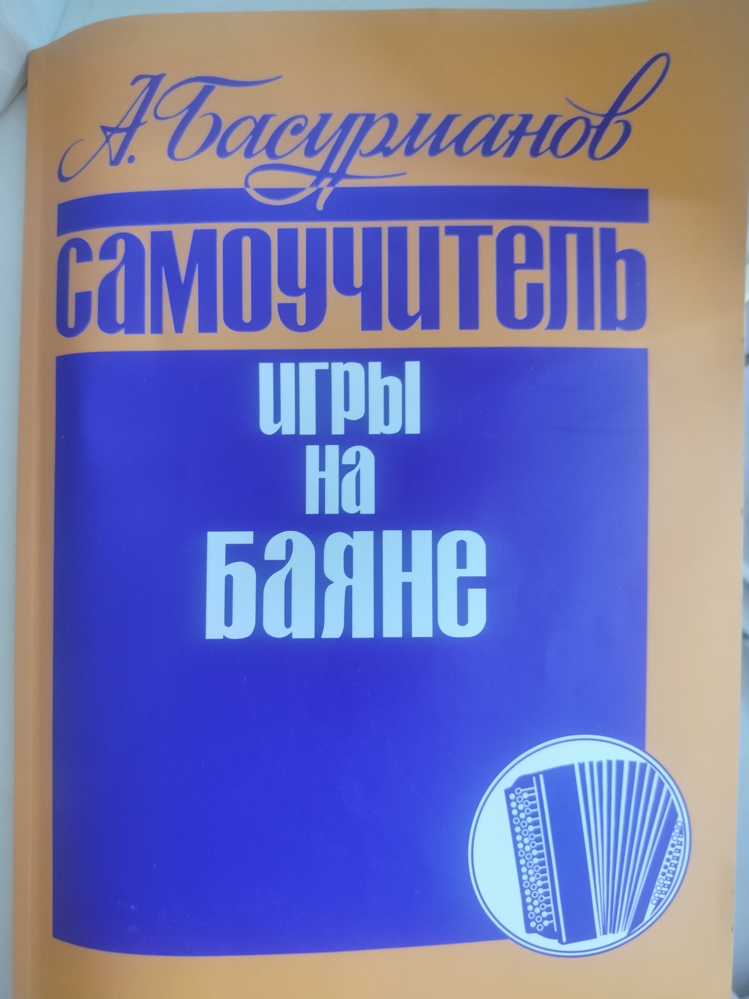 The image shows the cover of a book with a title in Russian. The title appears to be "Самоучитель игры на баяне," which translates to "Self-teacher of accordion playing." The author's name, "А. Басурманов," is written in a cursive font at the top. The book cover is orange with a blue rectangular area containing the title in white font. In the lower right corner, there is a circular emblem with an illustration of an accordion. The image is a flat, well-lit, indoor photograph. There is no indication of the location in Novosibirsk, Russia, in the image itself.