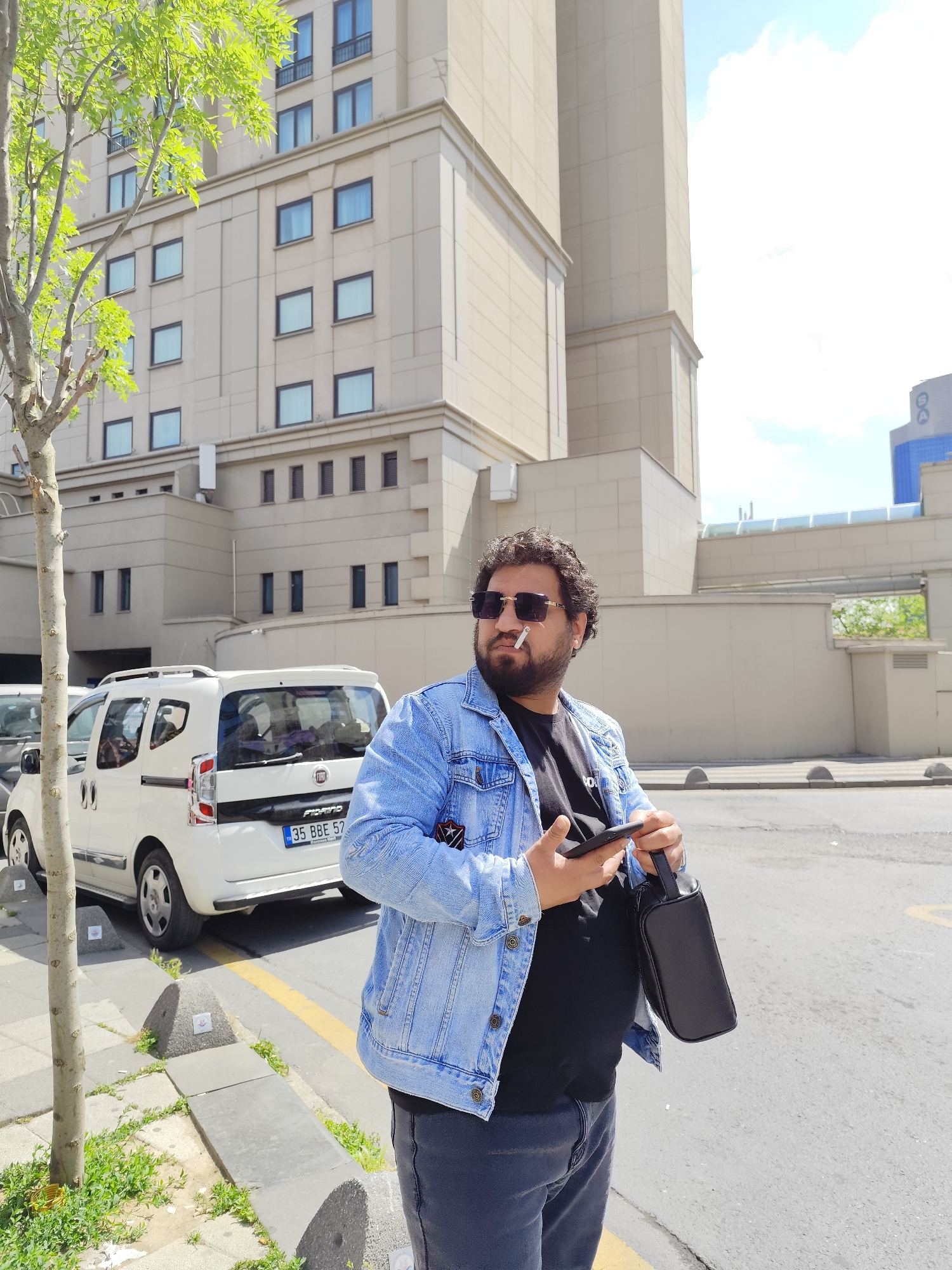 A man stands on a city sidewalk, holding a cell phone and a black bag. He is wearing sunglasses and a denim jacket, and smoking a cigarette. Behind him is a white vehicle parked near a building with multiple windows. The street is marked with a yellow line. The location is Unknown, Unknown.