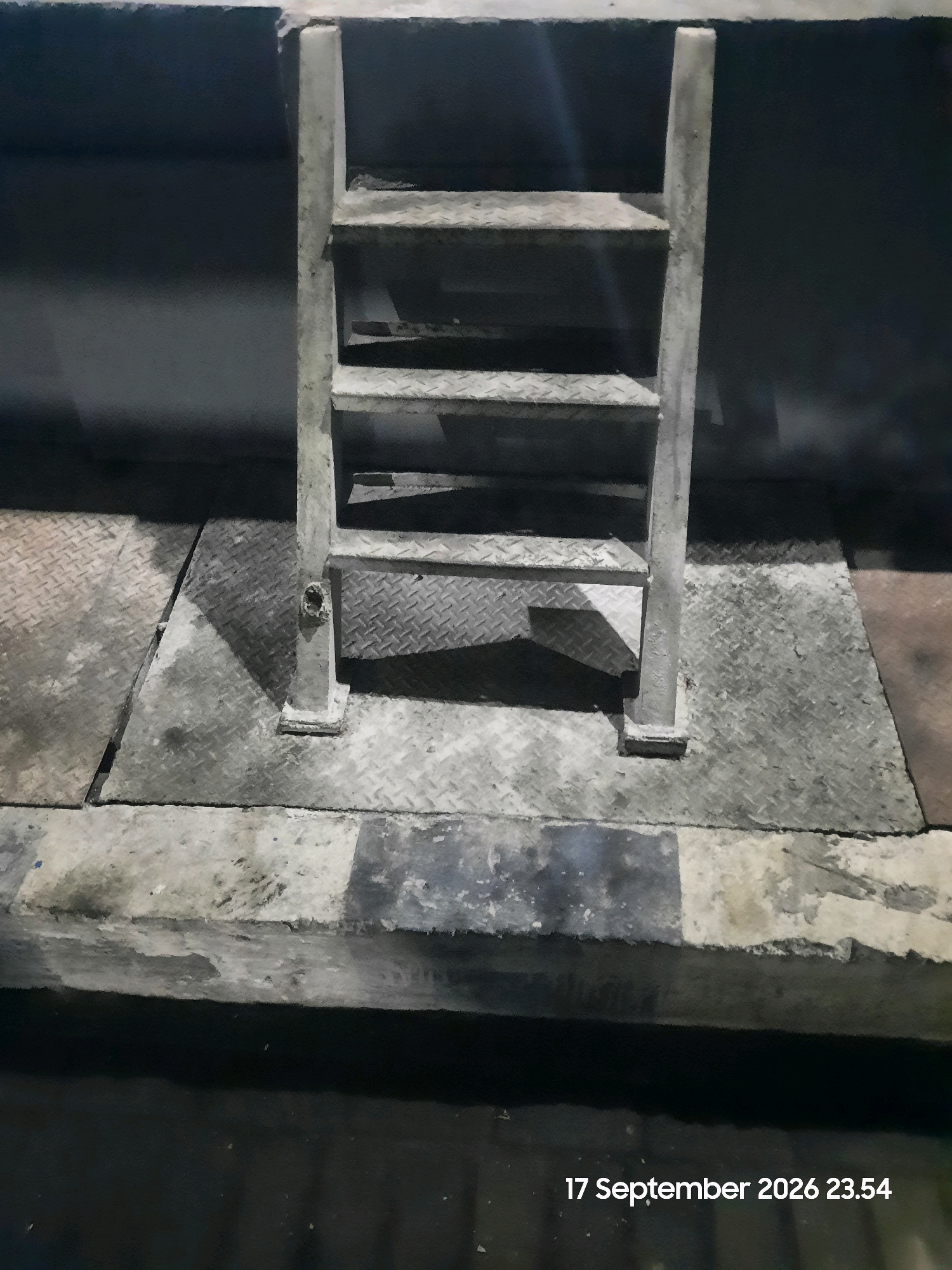 A metal ladder with diamond plate steps stands on a diamond plate platform in Gending, Indonesia. The ladder is painted a weathered, light gray and has two visible side rails and four steps. The platform is also made of diamond plate metal and is set on a concrete base with alternating dark and light gray painted sections. The background is a dark, textured wall. The scene is illuminated by artificial light, casting shadows from the ladder onto the platform. A timestamp in the lower right corner reads "17 September 2026 23.54".