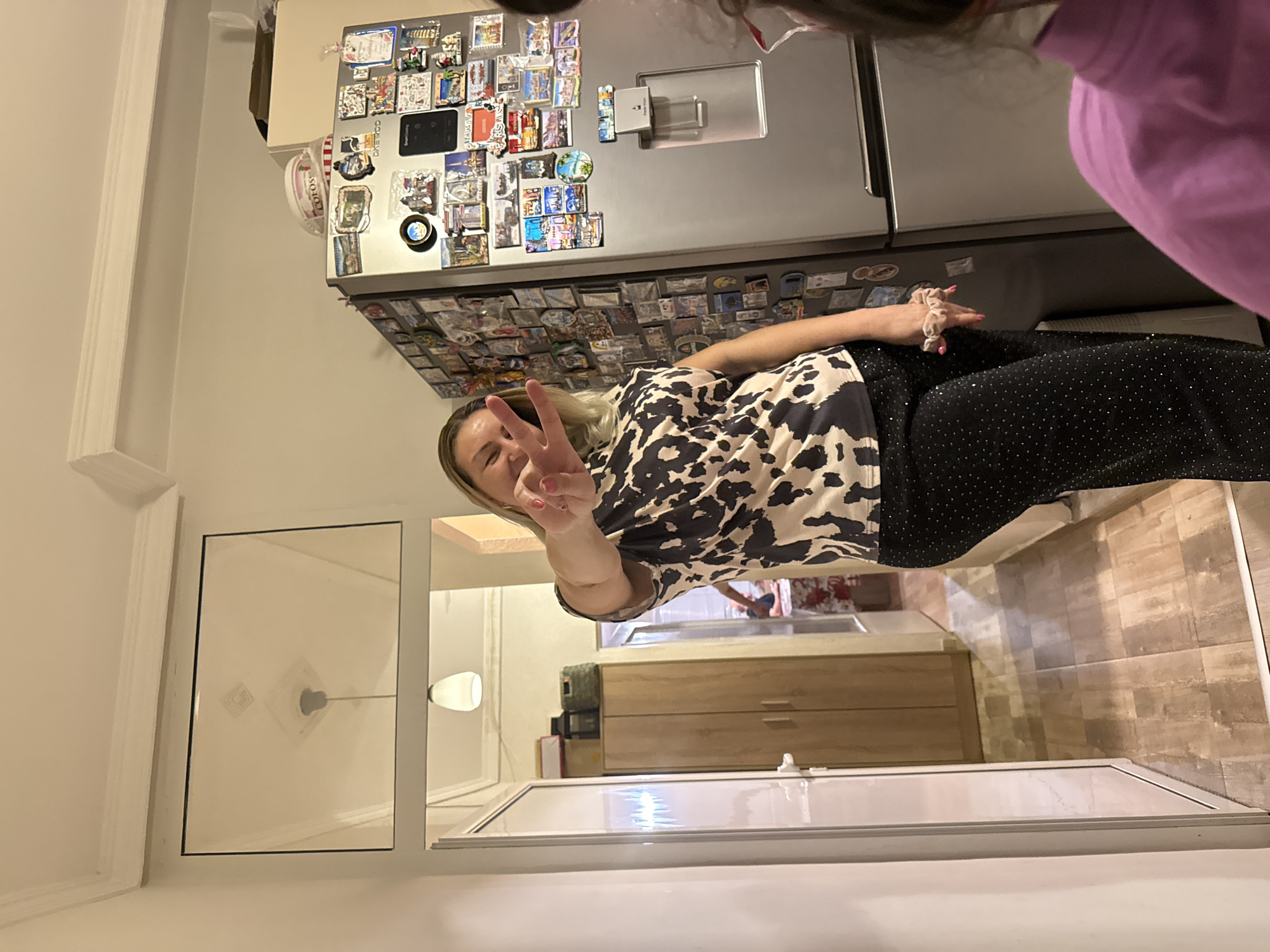 A woman with blonde hair is standing in a kitchen and giving a peace sign. She is wearing a black and white cow-print shirt and black pants with sequins. Behind her is a refrigerator covered in magnets. In the background, there is a hallway leading to another room with a light-colored countertop. A wooden cabinet is visible in the hallway. The location is likely a home kitchen, but the city of Stara Zagora, Bulgaria, cannot be verified from the image.