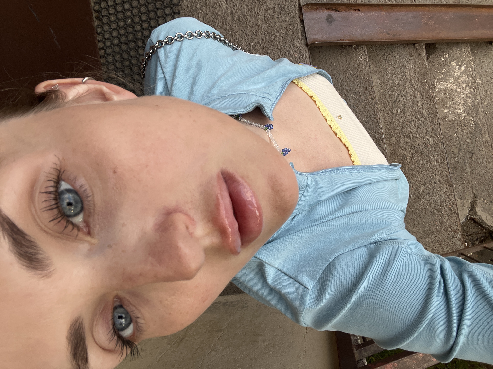 A close-up, high-angle, point-of-view shot features a young Caucasian woman's face as the primary subject. She has bright blue eyes, dark eyebrows, and fair skin with visible freckles. Her hair is light brown and pulled back, revealing her left ear, which has two small silver hoop earrings. She is wearing a light blue, ribbed, long-sleeved cropped shirt that is zipped partially down. Underneath, a white camisole with a yellow trim is visible. A delicate necklace with clear beads and small blue flower charms rests on her collarbone. The woman is looking directly into the camera with a neutral expression. The background consists of a textured, dark gray surface that appears to be concrete or pavement, with a rusty metal bar visible to the right and a dark brown textured surface above. The lighting suggests an outdoor setting, possibly during the day. The image is sharp and detailed, with a shallow depth of field that blurs the background.