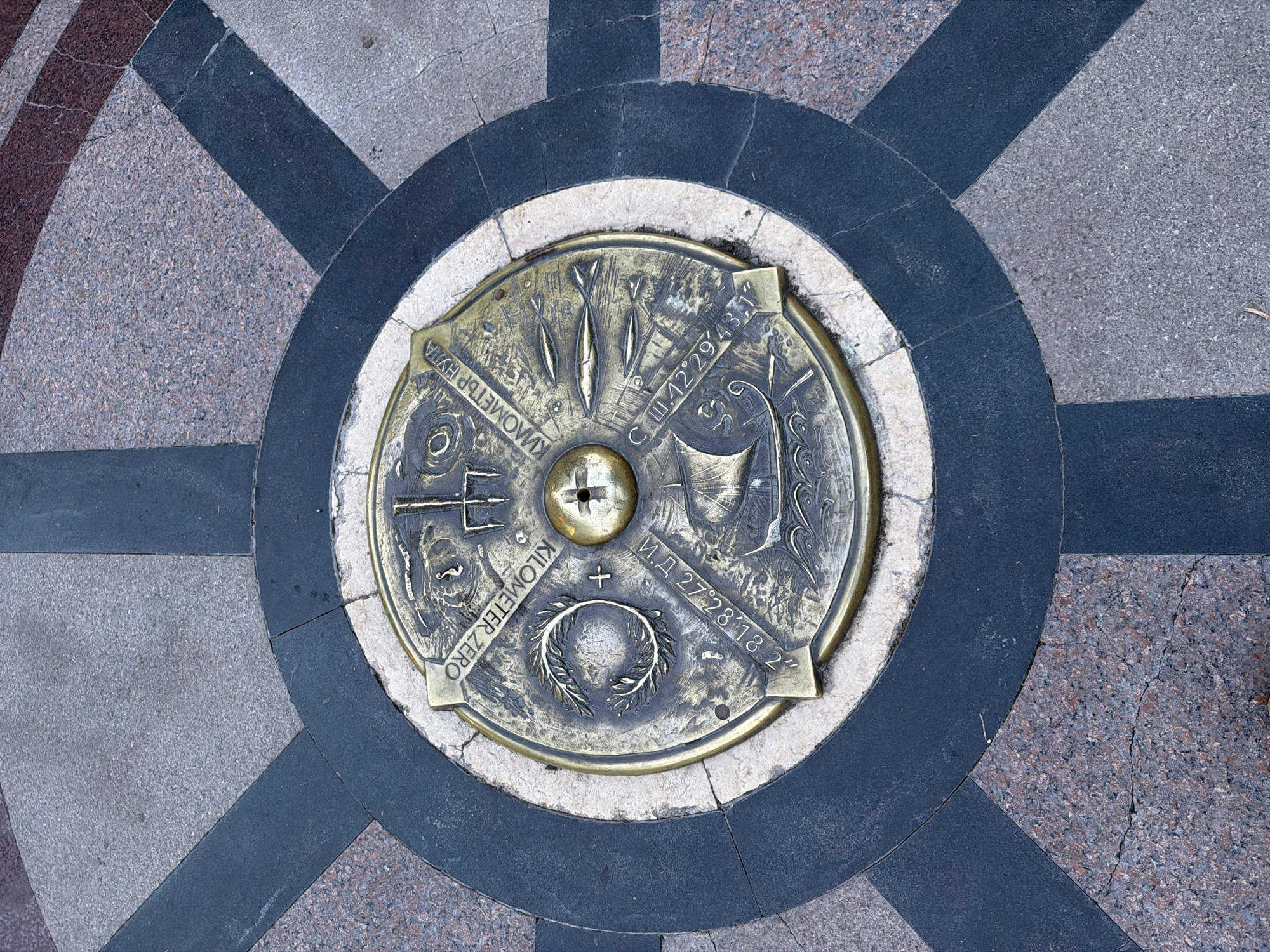 The image shows the "Kilometer Zero" marker in Burgas, Bulgaria. The marker is a circular bronze plate embedded in a circular pattern of black and light-colored stone tiles on the pavement. The bronze plate contains various symbols and inscriptions, including a compass design, geographic coordinates (С.Ш.42°29′43,1" and И.Д 27°28′18 2"), depictions of fish, and the phrase "KILOMETER ZERO" in both English and Cyrillic ("КИЛОМЕТЪР НУЛА"). The pattern of the stone tiles around the marker is designed with alternating dark and light stones, in a radiating pattern. The surrounding pavement consists of a speckled stone surface. There are no people visible in the image.