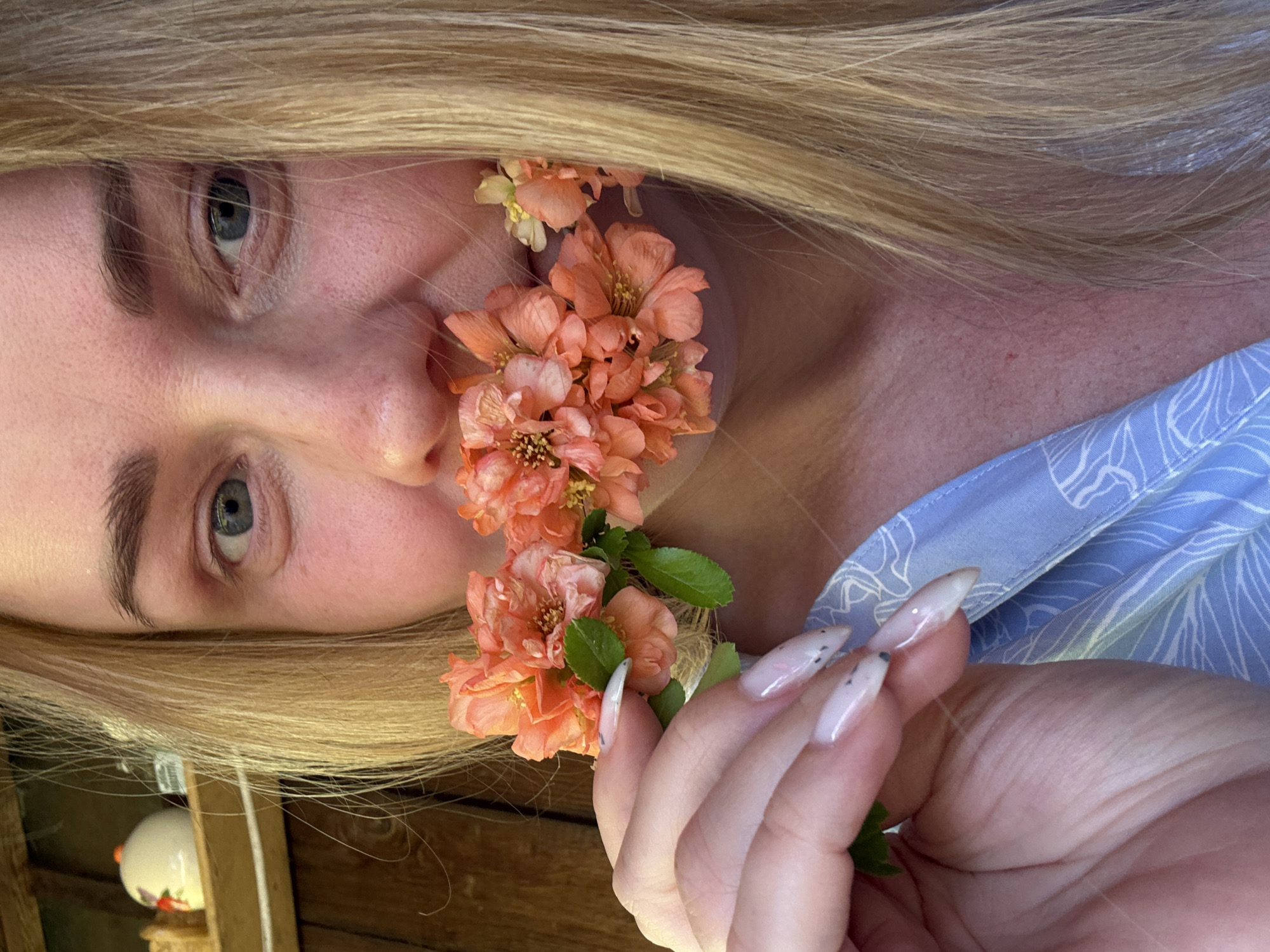 The image shows a Caucasian woman with blonde hair and blue eyes. Her face is partially obscured by a cluster of orange flowers. She is wearing a blue top with a white floral pattern. Her fingernails are long and decorated with a light pink base and dark spots. The background appears to be an outdoor setting with wood visible. The location is Chaikovsky, Russia.