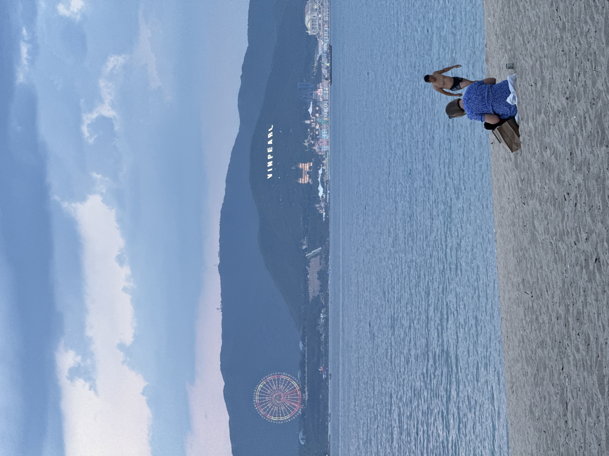 The image depicts a twilight scene at a beach in Nha Trang, Vietnam, overlooking a body of water with a distant city skyline and mountains. In the foreground, on a sandy beach, a woman wearing a blue polka-dot dress is seated, facing away from the camera, with a brown paper bag and a white object next to her. Behind her, a man in black swimming trunks is walking away from the water.
Across the water, a large Ferris wheel with illuminated red and yellow lights is visible on the left side of a mountain range. The word "VINPEARL" is illuminated in large white letters on the mountainside to the right of the Ferris wheel. Below the illuminated text, a colorful cityscape with various buildings, including a large arched structure and what appears to be a roller coaster, is illuminated against the darkening sky. The water is a calm blue, with gentle ripples. The sky is filled with cloudy, pale blue and gray hues. The overall atmosphere is serene, with the lights of the city and amusement park creating a vibrant contrast with the natural landscape.