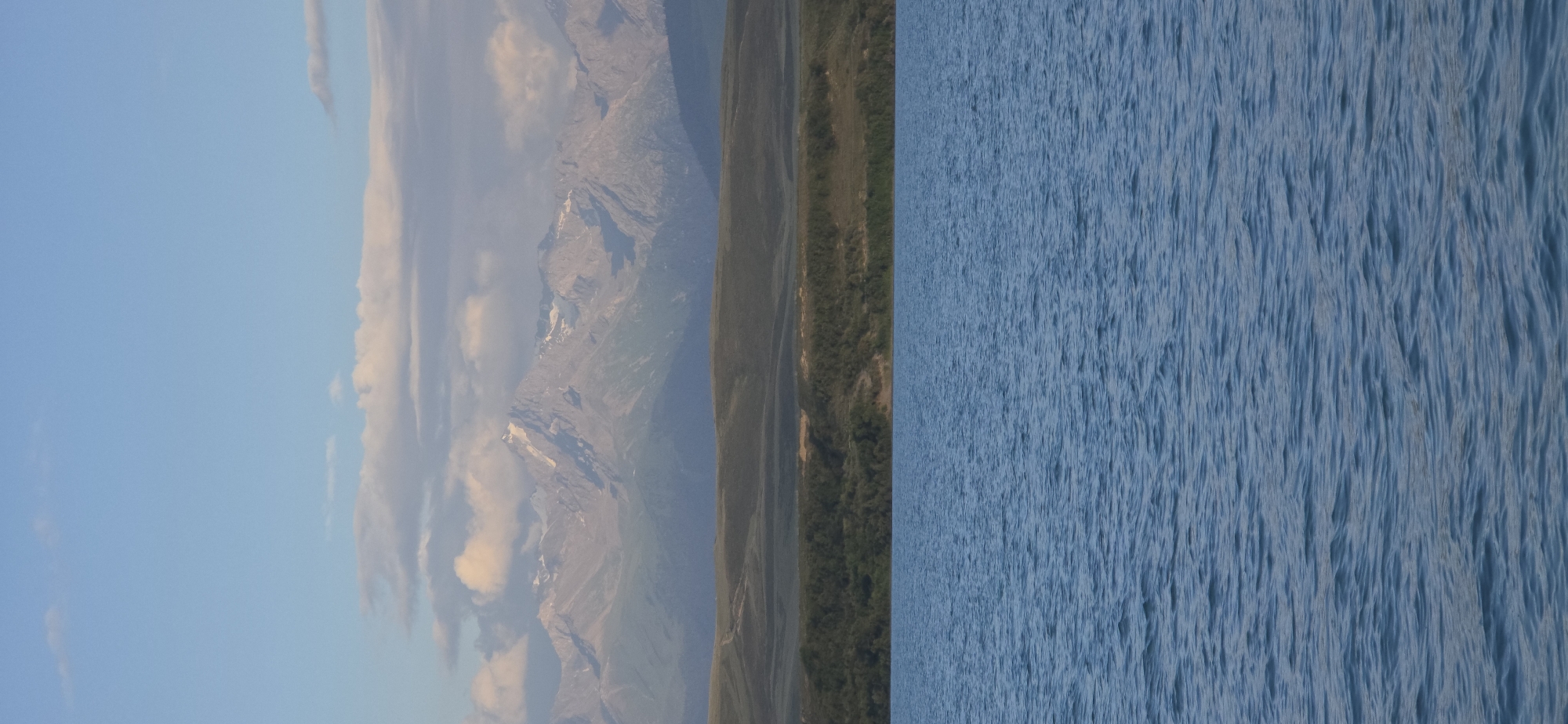 The image captures a serene natural landscape at Pristan'-Przheval'sk, Kyrgyzstan. In the foreground, a vast expanse of calm blue water stretches across the frame, its surface rippled by a gentle breeze. The water occupies the lower half of the image, creating a sense of depth and tranquility.
Above the water, a verdant green hillside rises, covered in dense vegetation that appears as a dark carpet. The hillside is textured with rolling contours and subtle variations in color, suggesting undulation and depth.
Further in the background, majestic mountains dominate the horizon. These peaks are rugged and imposing, with some of their summits capped with snow, gleaming white against the sky. The mountains exhibit a rugged, layered appearance, with sharp ridges and deep ravines, indicating a grand geological formation. The sky above is a pale blue, with scattered wispy clouds that add a touch of softness to the scene. The light suggests it is either early morning or late afternoon, casting a gentle glow over the landscape.
There are no people or man-made structures visible in the image, emphasizing the raw, untamed beauty of the natural environment. The overall impression is one of peace and grandeur, a moment frozen in time showcasing the natural splendor of the region.