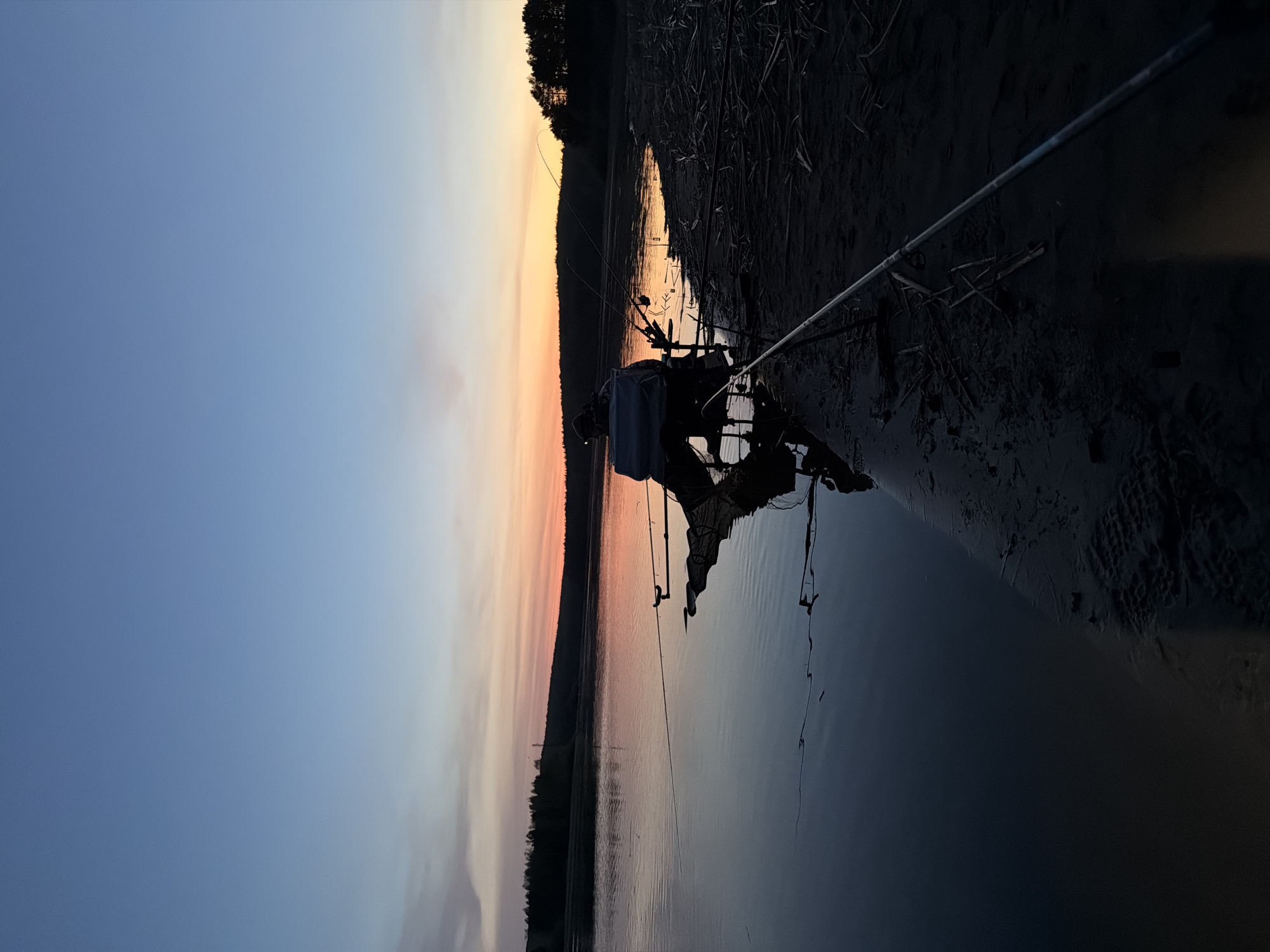 The image depicts a shoreline scene at dusk. The sky transitions from blue to orange, indicating either sunrise or sunset. A line of trees is silhouetted against the horizon. Fishing equipment, including rods, a chair, and a bag, are positioned on the muddy bank, extending into the water. The water is dark and reflective, mirroring the sky. The specific location in Chaykovsky, Russia, cannot be determined from the image alone.