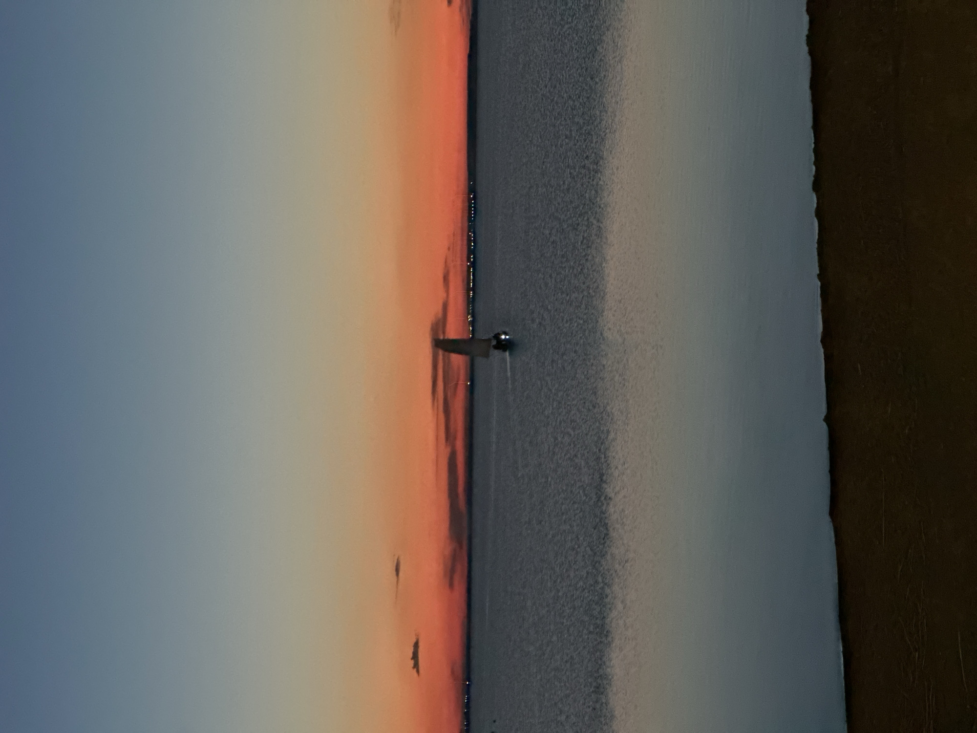 The image captures a tranquil scene of a single sailboat navigating calm waters under a twilight sky. The sky transitions from a deep blue at the top to a vibrant orange and red horizon, suggesting either sunrise or sunset. Wispy clouds are visible against the colorful sky. The water reflects the sky's hues, creating a serene mirror effect. In the distance, a faint line of lights indicates a shoreline or a populated area. The foreground is occupied by a dark, sandy embankment, possibly a beach or riverbank. The overall atmosphere is peaceful and serene, evoking a sense of calm. The location is identified as Lake Borovoye in Kokshetau, Kazakhstan, a popular resort area known for its natural beauty and recreational activities, including sailing.
