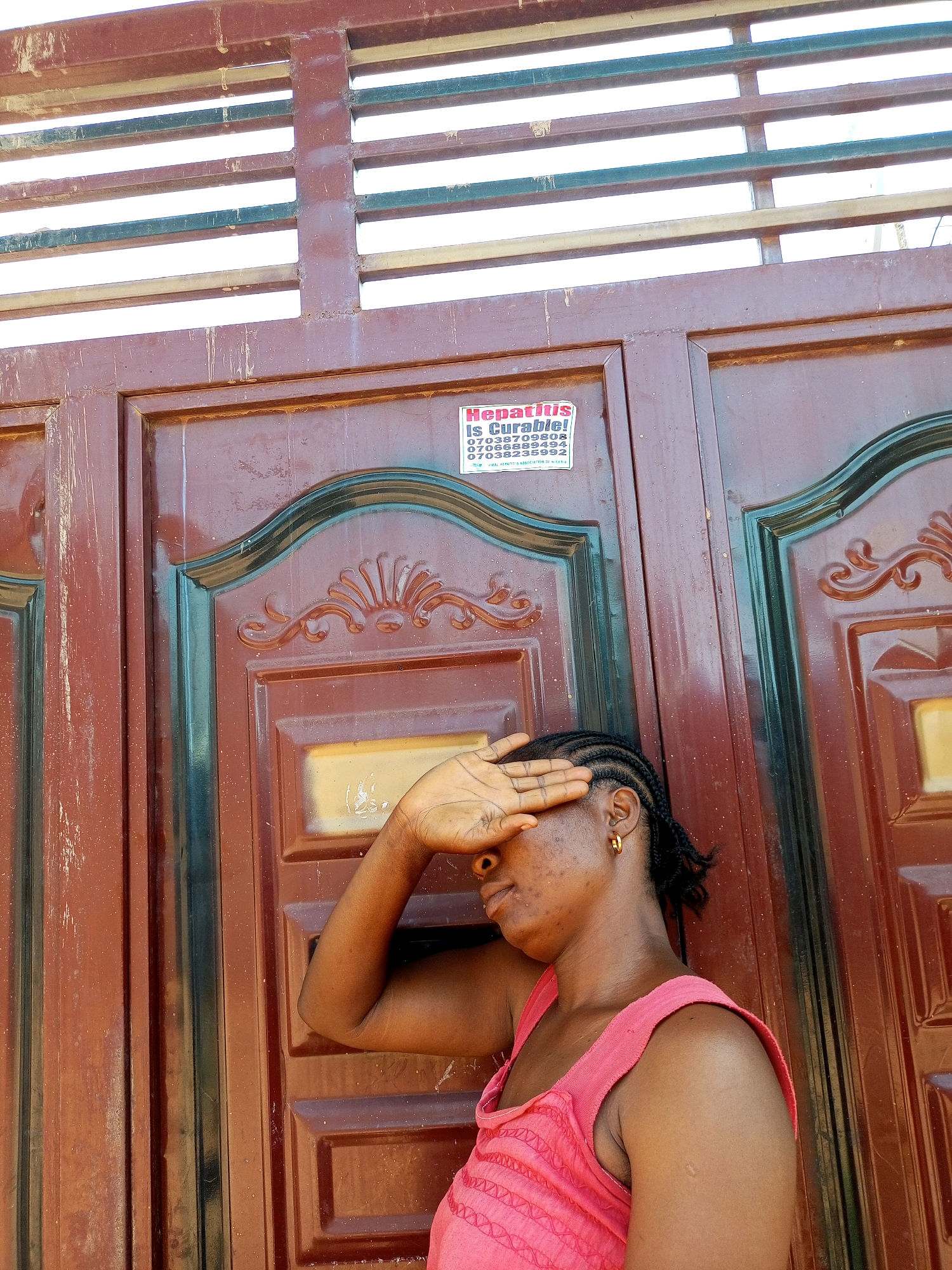 The image shows a medium shot of a woman standing in front of a closed, dark red metal gate with horizontal slats at the top. The woman is wearing a pink sleeveless top and has her right hand covering her eyes. She has braided hair and is wearing a gold hoop earring. A sticker with the text "HEPATITIS IS CURABLE!" and contact numbers is visible on the gate. The environment appears to be outdoors, with a bright sky visible through the gate's slats. The gate is adorned with decorative carvings. The context of Tempure, Nigeria, is not visually evident in the image itself, as no specific landmarks or identifying features of that location are present.