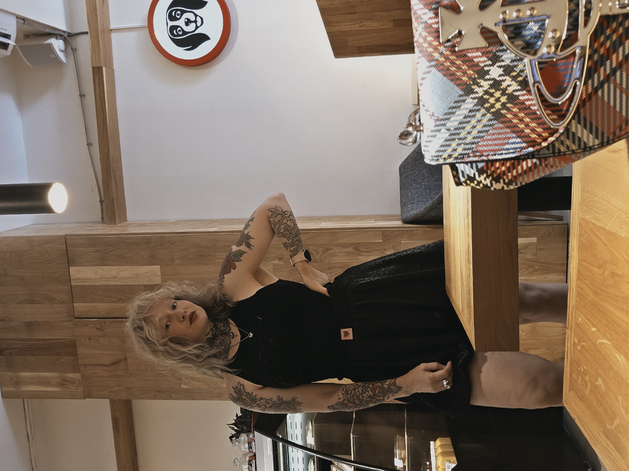 A blonde woman with voluminous curly hair and extensive floral tattoos on her arms and neck stands in what appears to be a cafe. She is wearing a black sleeveless top and a black skirt. Her left hand is on her hip, and her right hand is touching her hip. On the wall behind her, a circular sign with a dog's face is visible. In the foreground, a table with a plaid handbag adorned with a metallic orb and cross logo rests on it. The environment suggests an indoor setting with wooden paneling and furniture. The location is not specified as Saint Petersburg, Russia.