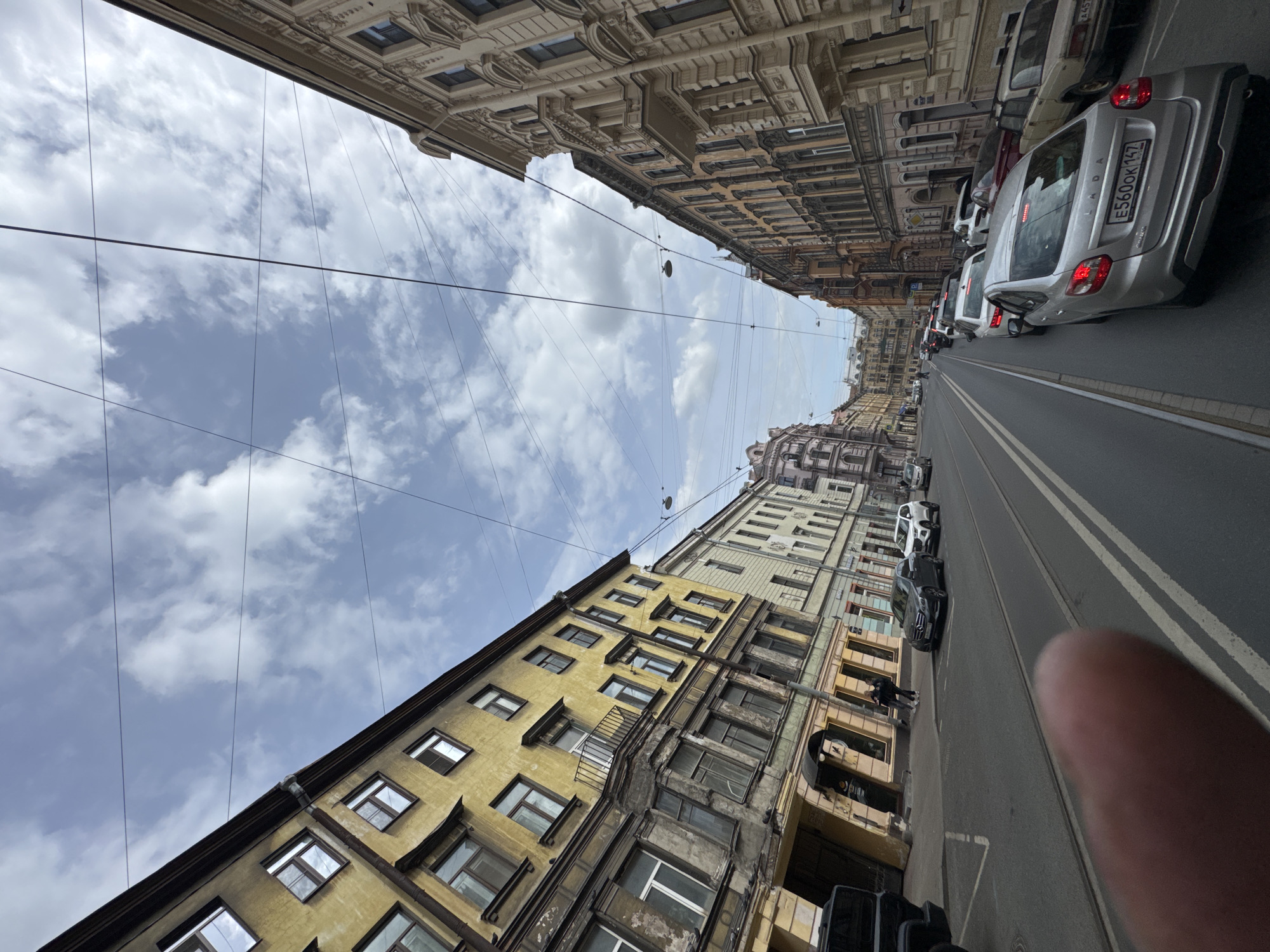 An eye-level street scene taken in Saint Petersburg, Russia, looking down a street toward buildings on both sides. The buildings are varied in color and architectural style, ranging from tan to yellow to brown, and are multi-storied. Overhead power lines run between the buildings. Cars are parked along the side of the street to the right, including a silver Lada with a visible license plate number E560OK142. Two people are standing on the sidewalk, and additional cars are further down the street. The sky is visible above the buildings and is partly cloudy with a mix of blue and white. A thumb is partially visible at the bottom right corner of the frame.