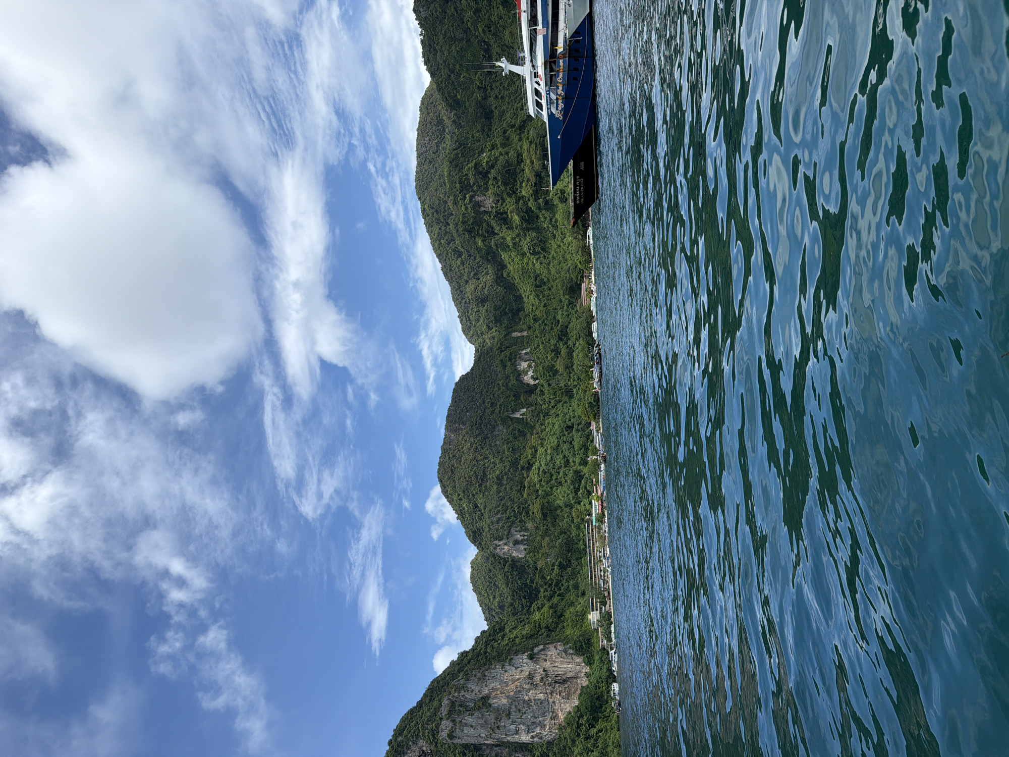A wide shot captures the turquoise waters of Ao Nang, Thailand, with lush green islands rising dramatically in the background. The water is choppy, reflecting the bright sky and scattered white clouds. Several boats are anchored in the bay, including a large blue and white cruise ship with the words "Royal Jet Cruise" visible on its side. A beach resort with low-rise buildings is nestled along the shoreline at the base of the islands. The steep, rocky faces of the islands are covered in dense vegetation, with some sheer cliffs visible. The image depicts a serene tropical scene.