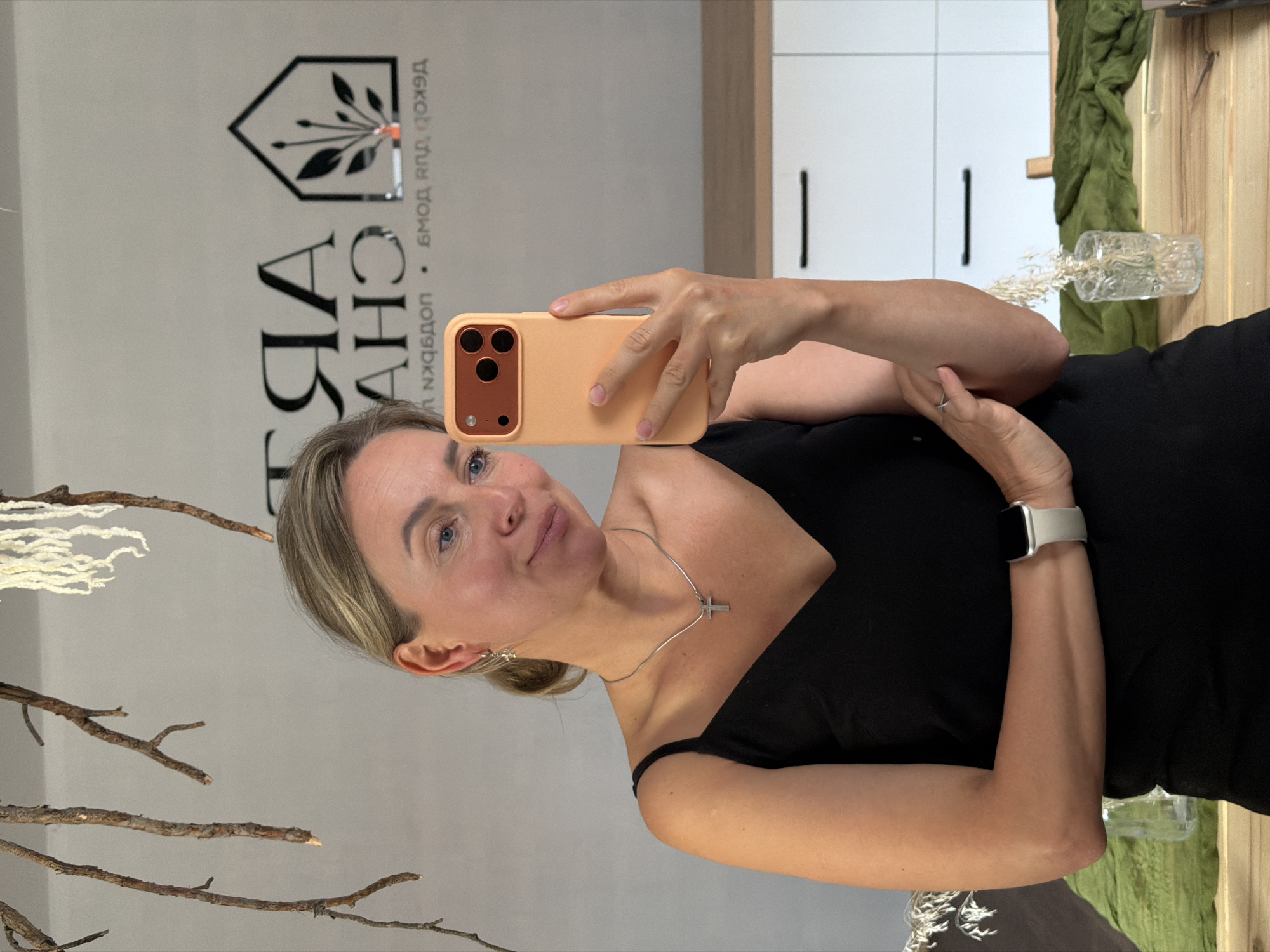 A woman with blonde hair, wearing a black camisole and a silver cross necklace, takes a mirror selfie in what appears to be an apartment in Chaikovsky, Russia. She is holding an orange phone in her right hand and her left arm is bent with her hand resting on her other arm. A white Apple Watch is visible on her left wrist. In the background, a mirror reflects a logo with text that reads "TRA AHG" and a house icon with plants. The environment includes wooden branches hanging from the ceiling and a dresser with white drawers.