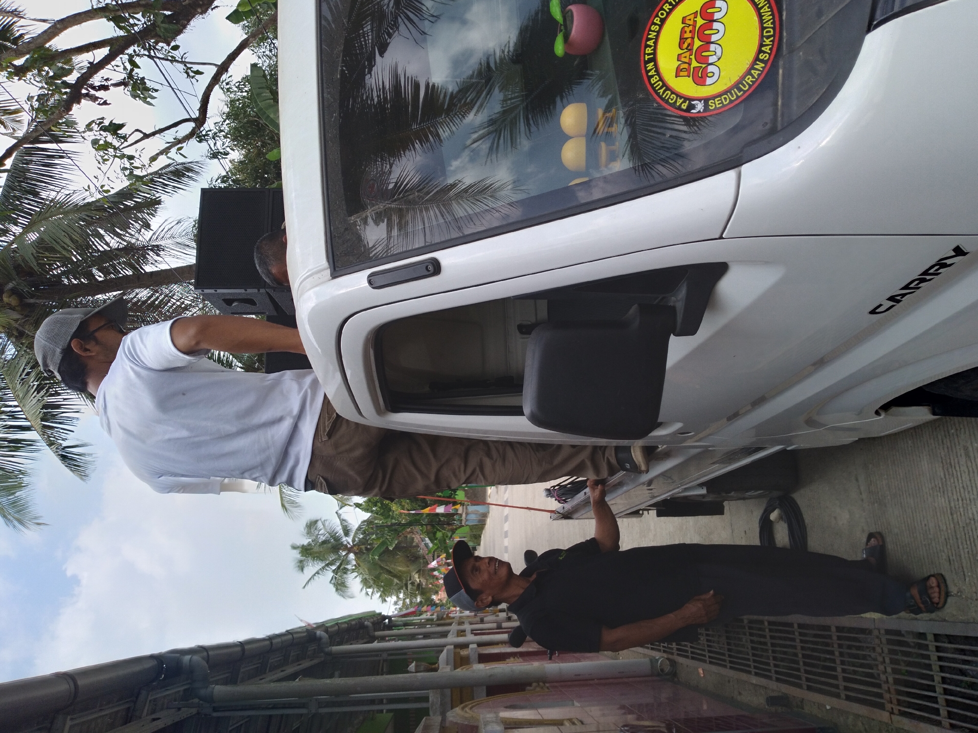 In Pandak, Indonesia, a man wearing a black polo shirt and dark pants stands on a concrete sidewalk next to a white pickup truck. He is looking to his left and smiling. Another man wearing a white t-shirt and brown pants is standing on the back of the pickup truck, leaning against a large black speaker. The truck has a sticker on the windshield with text that includes "DASRA 6000" and "SEDULURAN SAKDAWAN". The background shows a street lined with buildings and palm trees, with a cloudy sky overhead. There are also flags and decorative elements visible along the street.